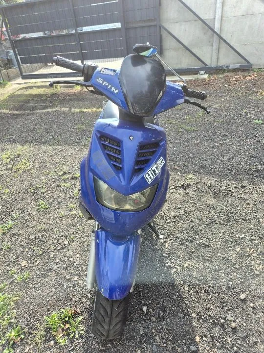 Niebieski skuter marki Peugeot Spin zaparkowany na żwirowym podłożu. Pojazd jest widoczny od przodu, posiada czarną owiewkę nad reflektorem oraz napis 'SPIN' na kierownicy. Na przedniej obudowie znajduje się naklejka z literami 'BT'. W tle widoczna jest ciemna brama i betonowy mur.