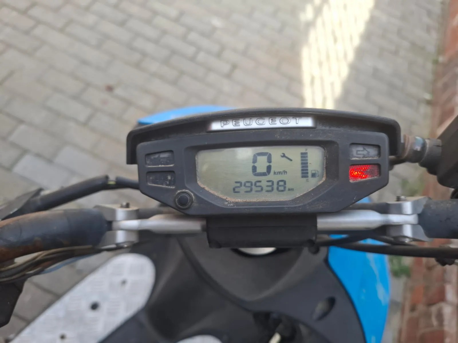 Zbliżenie na deskę rozdzielczą skutera marki Peugeot. Cyfrowy wyświetlacz pokazuje prędkość 0 km/h oraz przebieg 29538 km. Obok liczb widoczne są ikony klucza serwisowego i poziomu paliwa. Po prawej stronie znajduje się czerwona kontrolka. Całość osadzona jest w czarnej obudowie zamontowanej na srebrnym kierownicy.