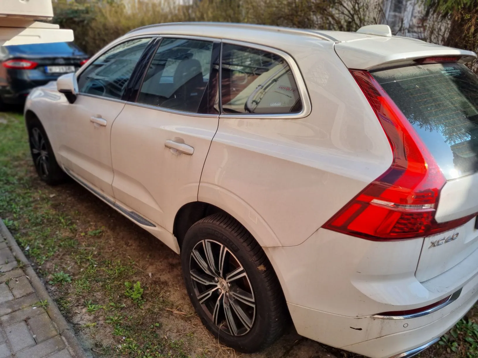 Biały samochód typu SUV marki Volvo model XC60 zaparkowany na trawie obok brukowanej ścieżki. Widoczne są czerwone światła tylne, srebrne klamki drzwi oraz czarne felgi. W tle częściowo widoczny czarny samochód i zarośla. Na tylnym zderzaku widnieje napis XC60.