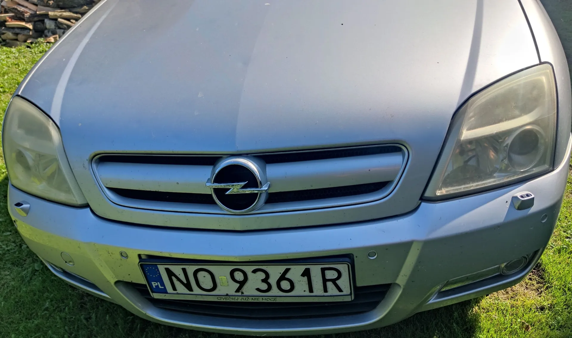 Zbliżenie na przód srebrnego samochodu marki Opel. Widoczny jest grill z logo producenta, reflektory oraz polska tablica rejestracyjna o numerze NO 9361R. Poniżej numeru rejestracyjnego znajduje się mały napis: 'SZWENIEJ JUŻ NIE MOGĘ'.
