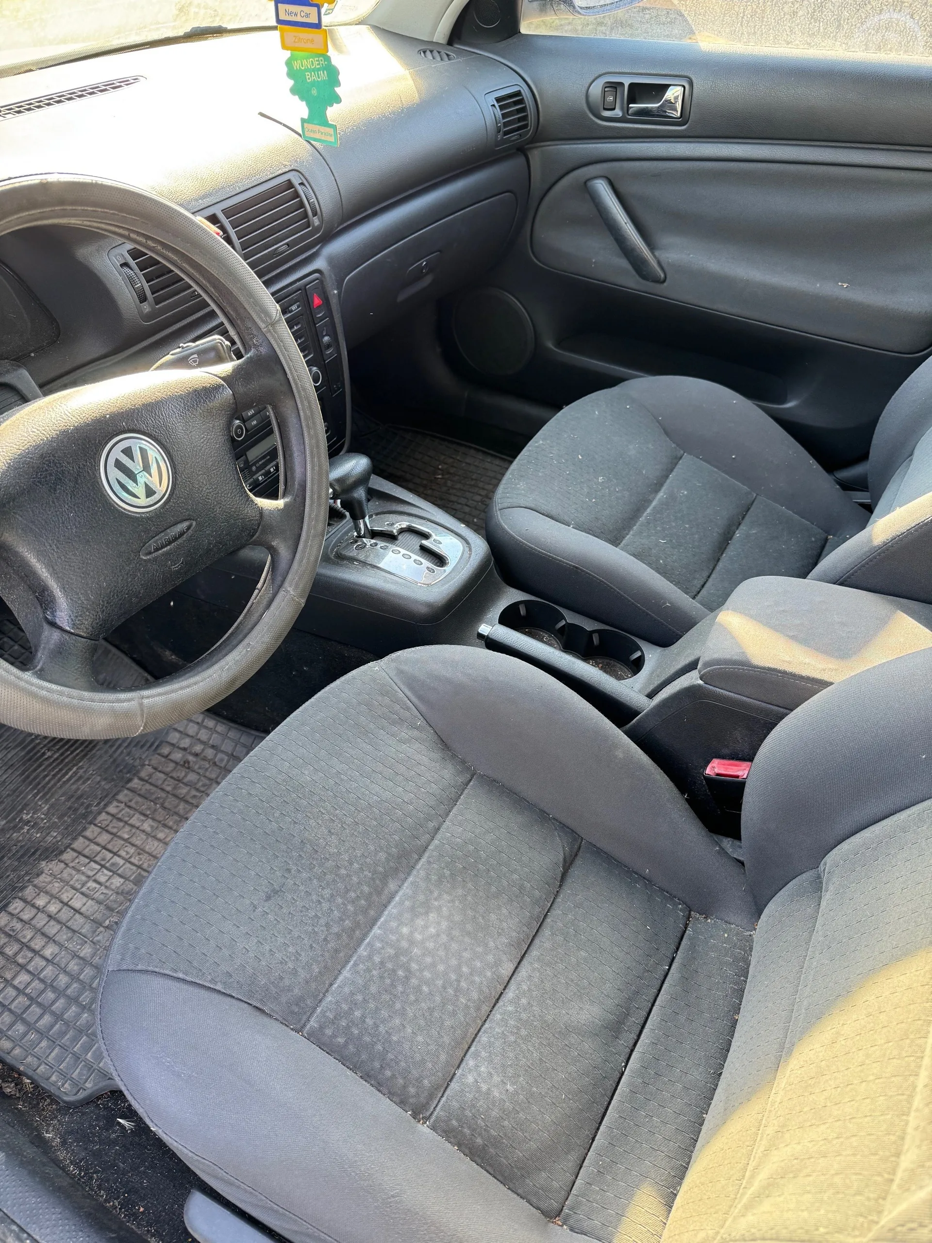 Wnętrze samochodu marki Volkswagen z czarną tapicerką siedzeń i ciemnoszarym panelem deski rozdzielczej. Widoczne kierownica z logo VW, dźwignia zmiany biegów, podłokietnik z uchwytem na kubki oraz czarne dywaniki podłogowe. Na desce rozdzielczej znajdują się naklejki: niebieska z napisem 'New Car', żółta oraz zielona z napisem 'WUNDER-BAUM'. Siedzenia pokryte są lekkim brudem, a w tle widoczna jest czarna obudowa drzwi z uchwytem i otworem wentylacyjnym.