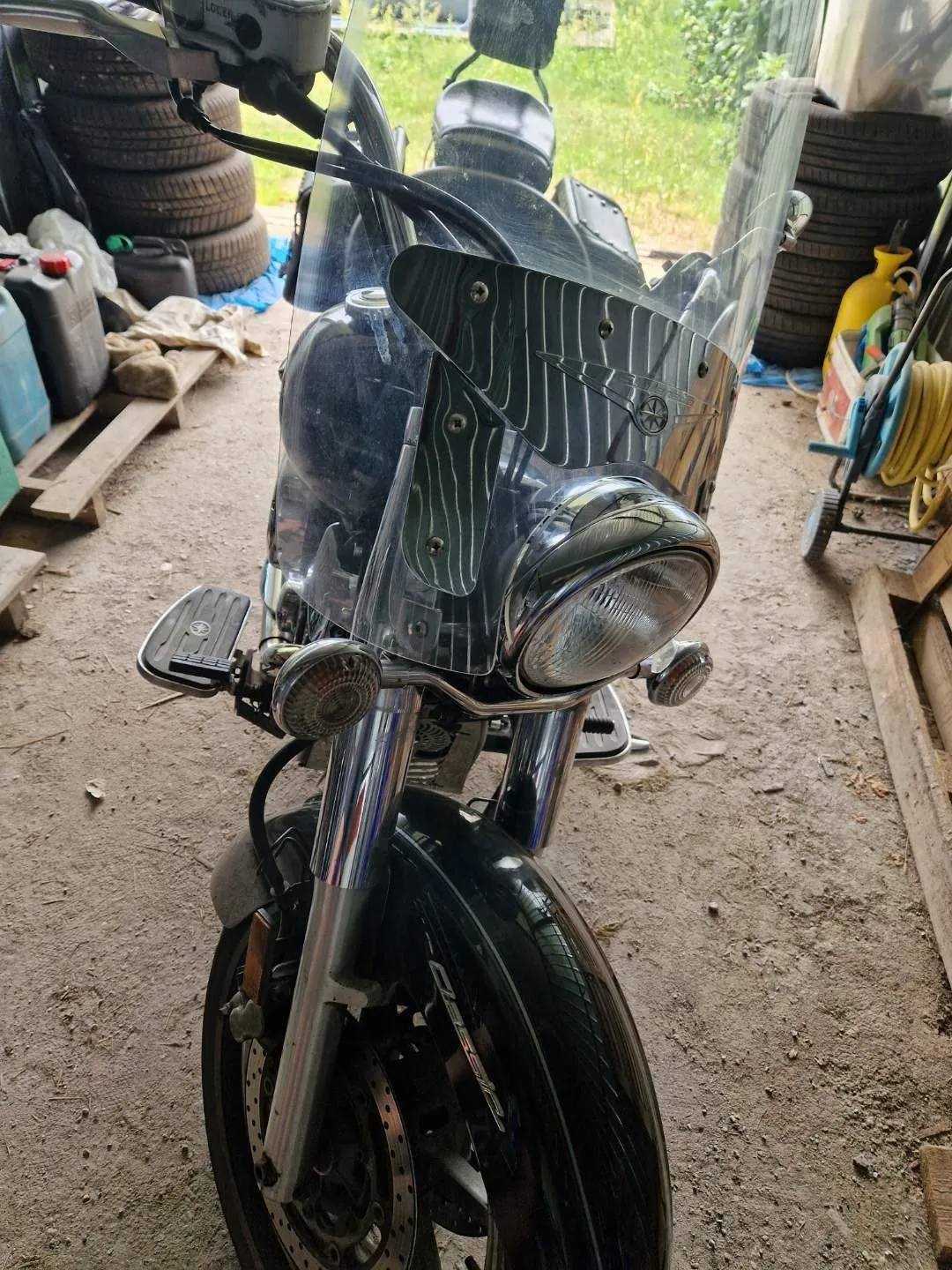 Zdjęcie przedstawia przód czarnego motocykla z dużą, przezroczystą szybą ochronną, na której widnieje ozdobny wzór w postaci zielonych linii. Motocykl posiada chromowane elementy, w tym przedni widelec oraz obudowę dużego, okrągłego reflektora. Widoczne są również mniejsze kierunkowskazy po obu stronach reflektora. Motocykl stoi w garażu lub warsztacie na betonowej posadzce, w tle widać stosy opon oraz kanistry.