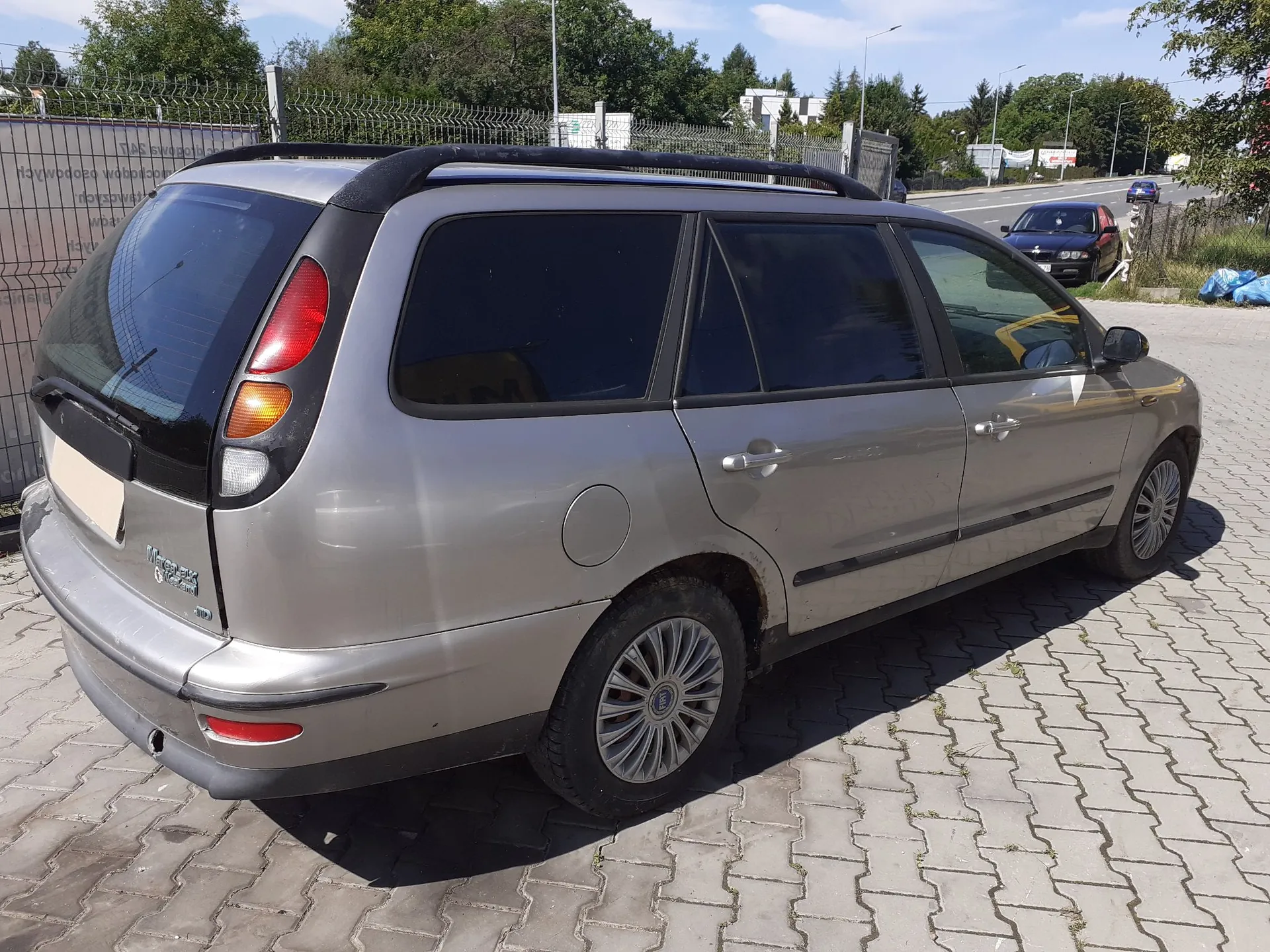 Srebrny samochód typu kombi zaparkowany tyłem i bokiem na kostce brukowej. Pojazd posiada czarne relingi dachowe, przyciemniane tylne szyby i wieloramienne felgi aluminiowe. W tle widoczny jest metalowy płot, drzewa oraz fragment drogi z innym samochodem.