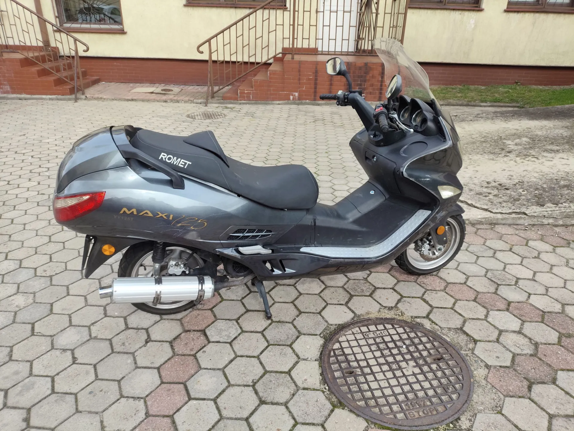 Szary skuter marki Romet, model Maxi 125, zaparkowany na kostce brukowej przed budynkiem. Pojazd ma czarną kanapę, srebrną rurę wydechową i wysoką szybę ochronną. Na boku skutera widnieją napisy 'ROMET' oraz 'MAXI 125'.