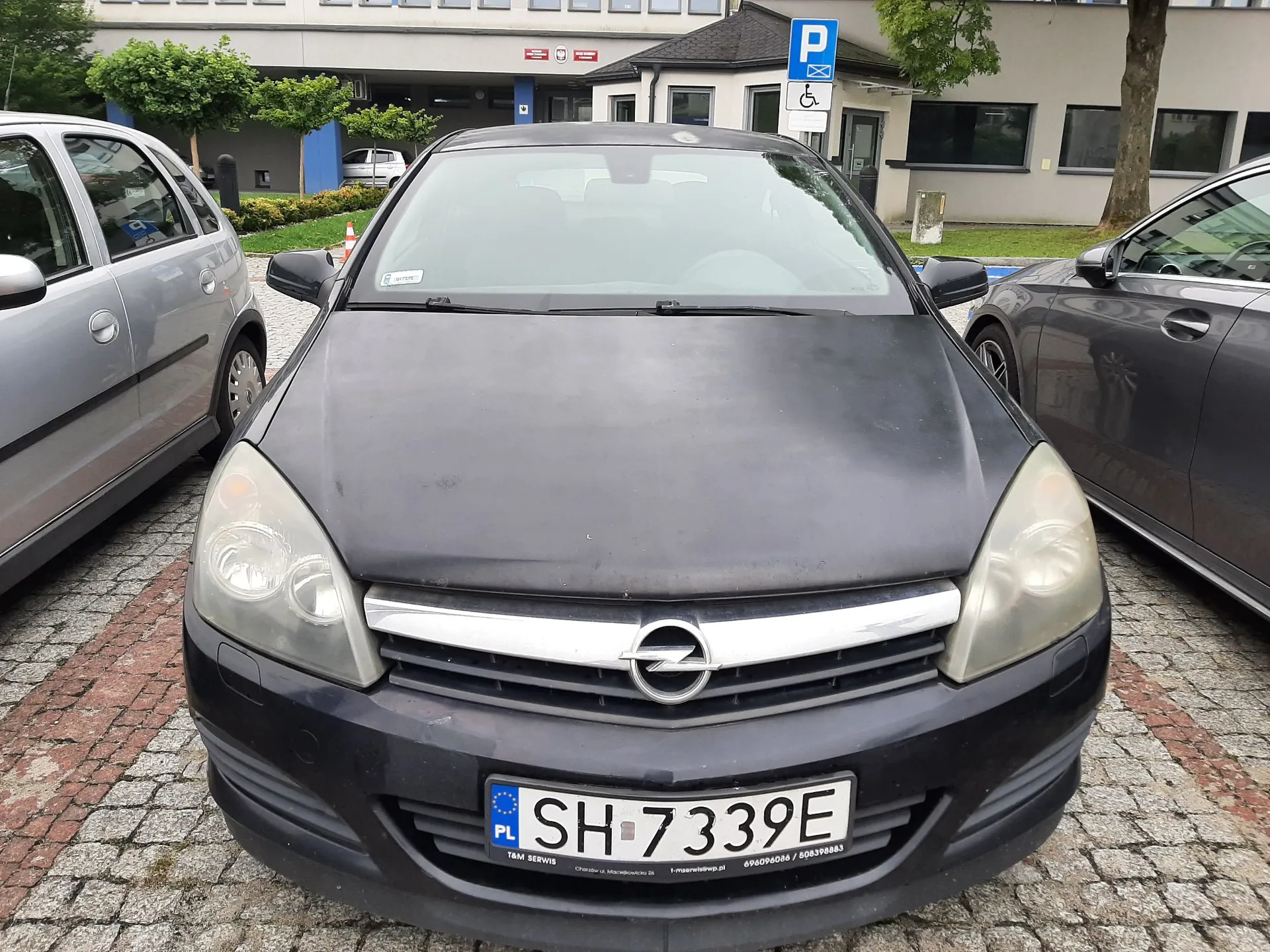 Zdjęcie przodu czarnego samochodu marki Opel zaparkowanego między dwoma innymi pojazdami na kostce brukowej. Samochód ma tablicę rejestracyjną o numerze SH 7339E. W tle widoczny jest budynek biurowy oraz znak parkingu dla osób niepełnosprawnych.