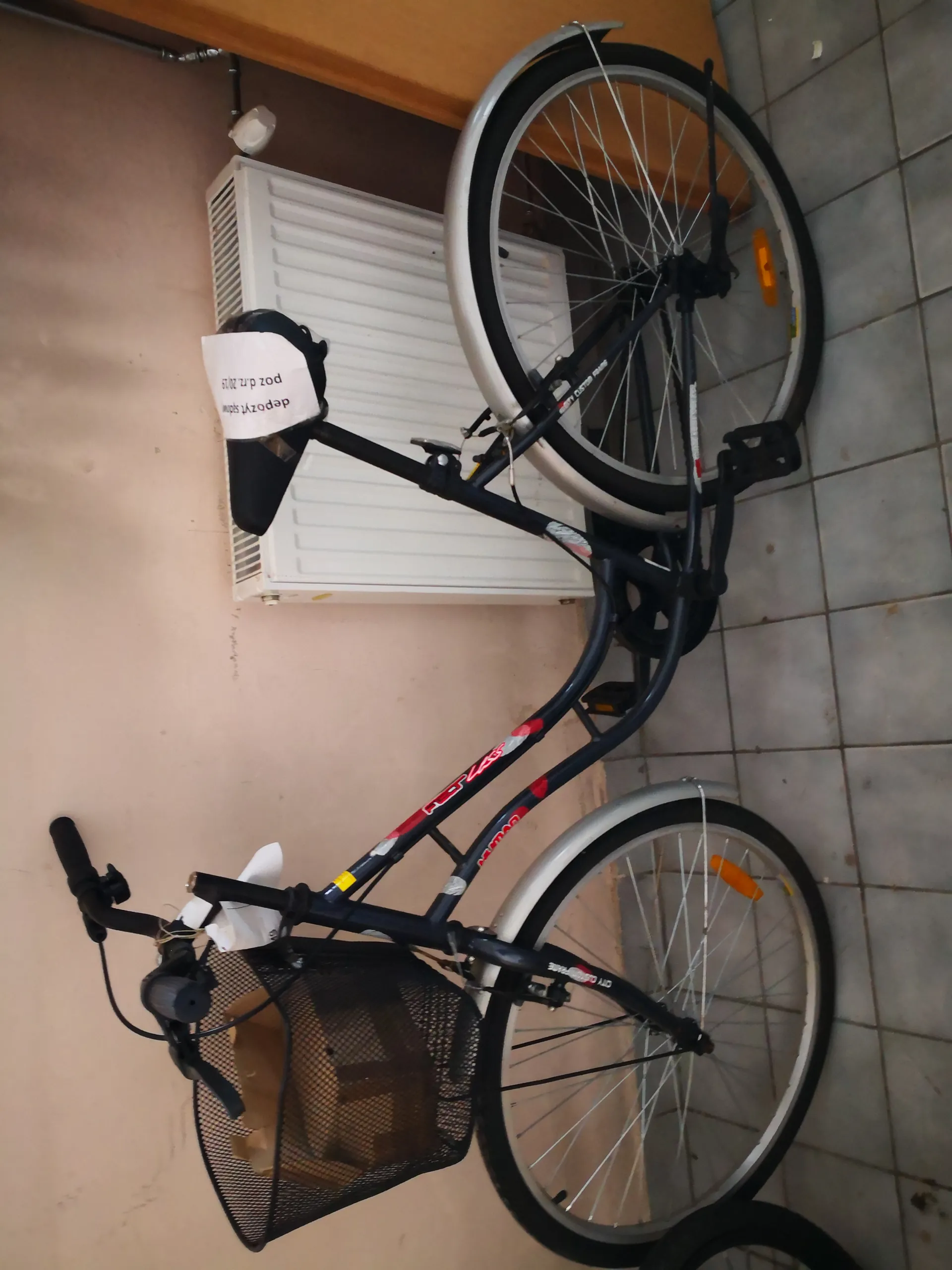 Ciemnoniebieski rower z czerwonymi akcentami, z czarnym siatkowym koszem na przednim kole zawierającym karton. Na kierownicy biała etykieta, na siodełku biała etykieta z tekstem 'pozdr...'. Rower oparty o beżową ścianę obok białego grzejnika. Podłoga wyłożona szarymi płytkami. Widoczne pomarańczowe odblaski na obu kołach.