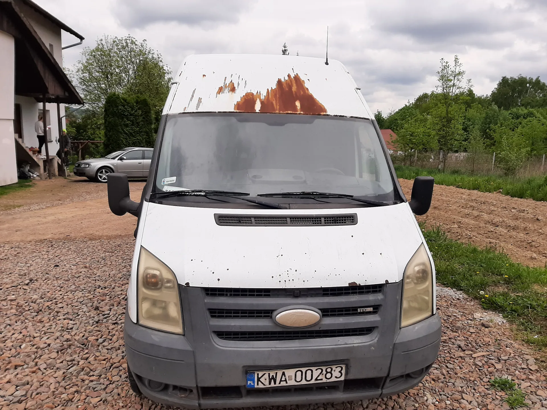 Przedni widok białego furgonu marki Ford z widoczną rdzą na dachu. Tablica rejestracyjna PL KWA-00283. Furgon stoi na żwirowym podjeździe. W tle dom z osobą stojącą przy wejściu, srebrny samochód osobowy, zielone krzewy i drzewa oraz chmurne niebo.