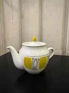 Biały czajnik z żółtymi okrągłymi wzorami i dekoracyjnym motywem na boku, umieszczony na ciemnej powierzchni. Tło stanowią jasne, pionowe panele.