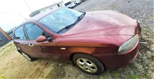 Czerwony samochód osobowy Alfa Romeo 156, zaparkowany na podwórku z żwirem i ziemią. Widoczne są lekkie uszkodzenia lakieru, cztery koła z metalowymi felgami, przednie reflektory oraz boczne lusterko. W tle budynki i częściowo widoczny inny pojazd. Samochód znajduje się na otwartym obszarze z nieutwardzonym podłożem.