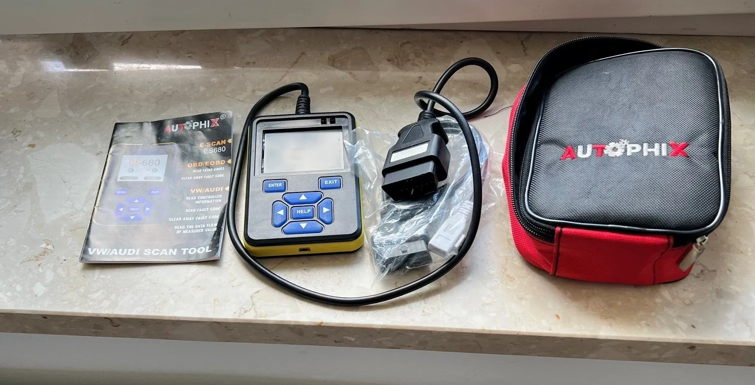 Na marmurowym blacie umieszczono zestaw narzędzi diagnostycznych samochodowych marki AUTOPHIX. Po lewej stronie znajduje się instrukcja z napisami 'AUTOPHIX', 'E-SCAN ES680', 'OBD/EOBD', 'VW/AUDI SCAN TOOL' oraz grafiką urządzenia. W centrum leży urządzenie z ekranem, przyciskami 'ENTER', 'EXIT', 'HELP' i kierunkowymi strzałkami, połączone czarnym kablem z czarnym złączem OBD-II. Obok, w przezroczystej folii, widoczne są dodatkowe akcesoria. Po prawej stronie znajduje się czerwono-czarna torba z zamkiem, na której widnieje czerwony napis 'AUTOPHIX'.