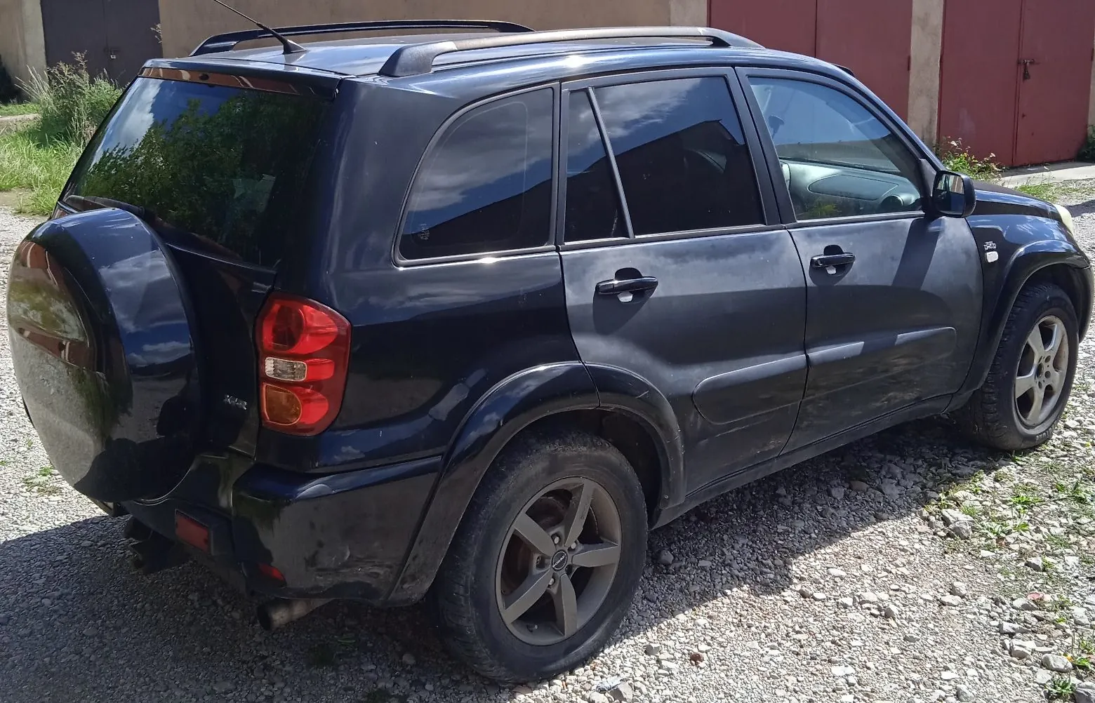 Czarny samochód typu SUV zaparkowany na żwirowym podłożu. Pojazd jest widoczny z tyłu i z boku, posiada zamontowane koło zapasowe na klapie bagażnika oraz relingi dachowe. W tle widoczny jest fragment budynku z czerwonymi drzwiami.