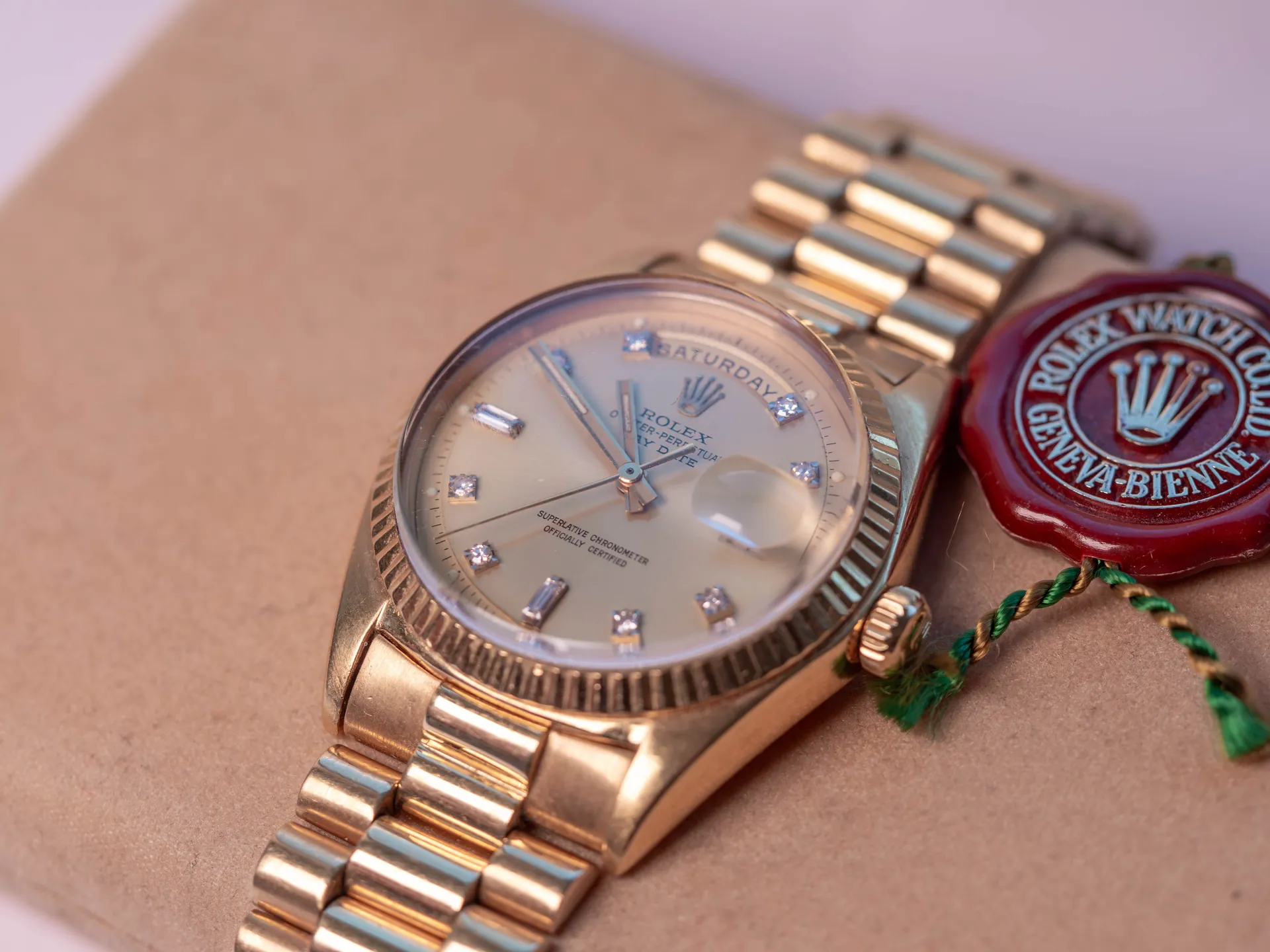 Złoty zegarek marki Rolex z bransoletą, leżący na brązowej teczce lub pudełku. Tarcza zegarka jest jasna z naniesionymi indeksami w formie małych kamieni i napisem 'ROLEX OYSTER PERPETUAL DAY-DATE'. Na tarczy widoczny jest dzień tygodnia 'SATURDAY'. Obok zegarka znajduje się czerwona lakowa pieczęć z logo Rolex i napisem 'ROLEX WATCH CO GENEVA-BIENNE'.
