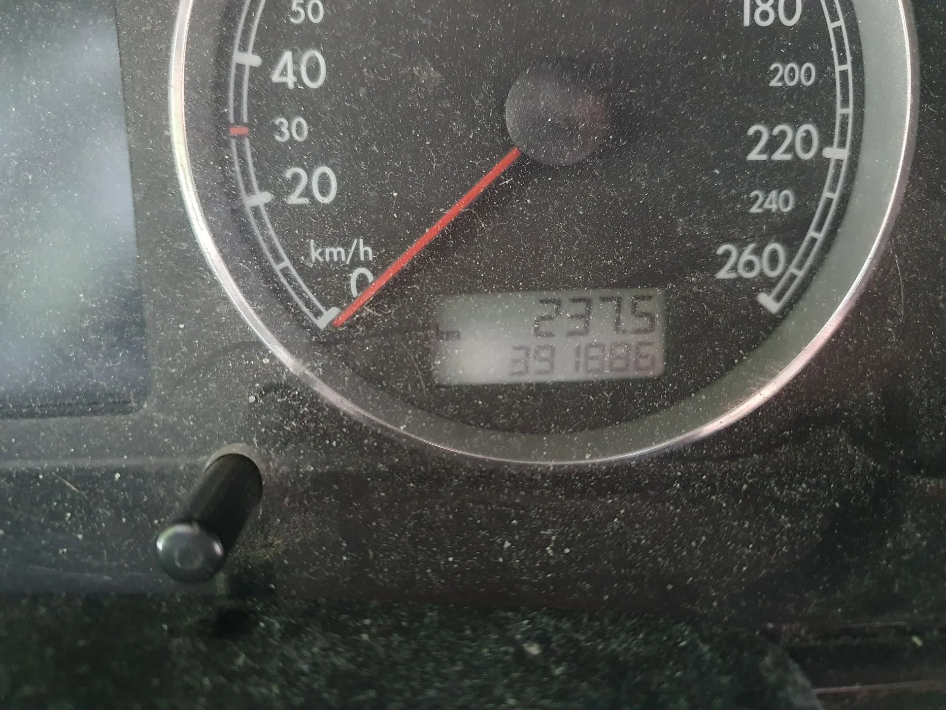 Zbliżenie na okrągły prędkościomierz w samochodzie. Skala prędkości sięga 260 km/h, a wskazówka znajduje się w pozycji zerowej. Na dole tarczy widoczny jest cyfrowy wyświetlacz z liczbami 237.5 oraz 391886. Powierzchnia deski rozdzielczej jest zakurzona.