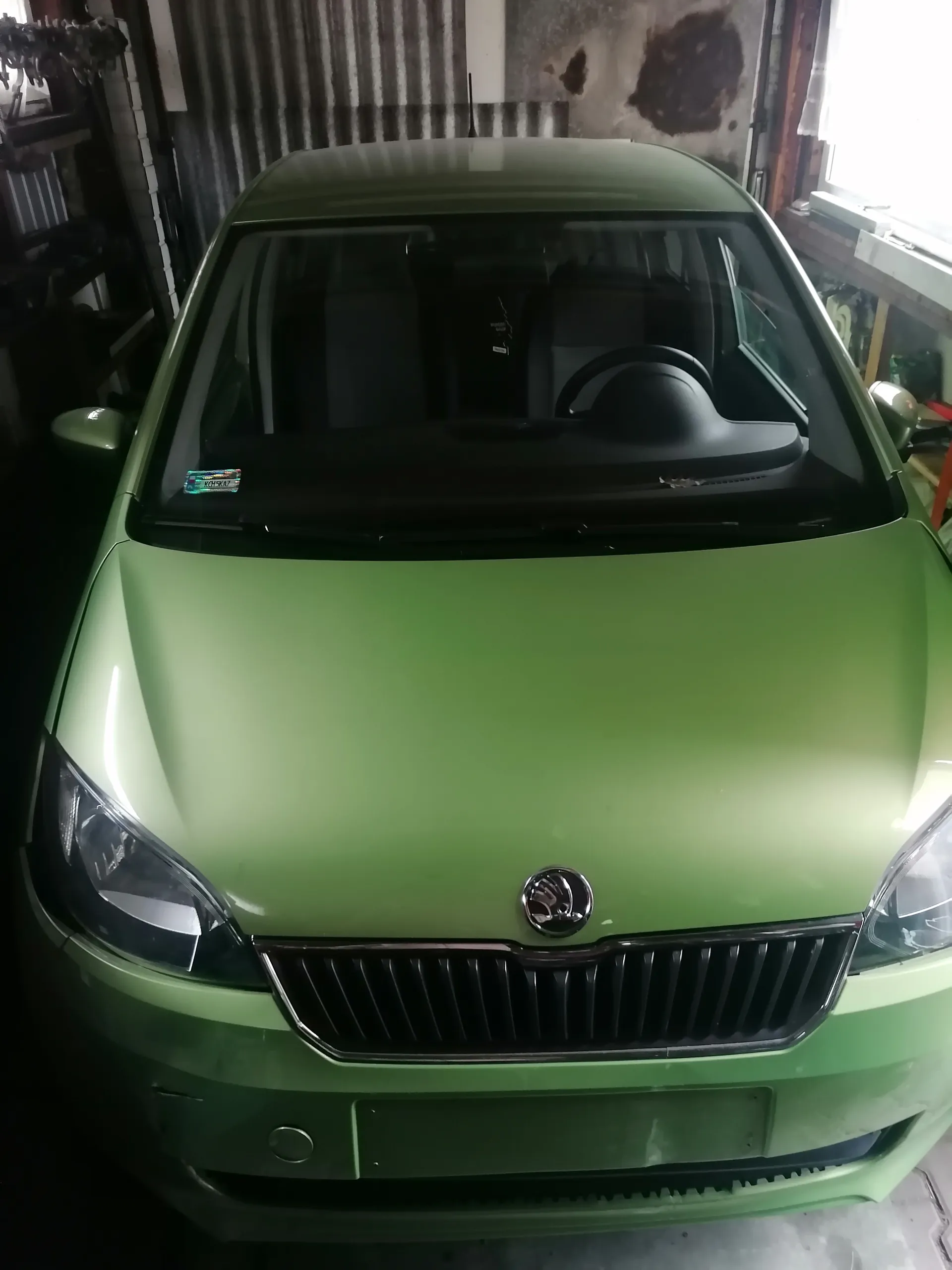 Zielony samochód marki Škoda zaparkowany w garażu. Widoczna przód pojazdu z logo na masce, reflektorami i atrapą chłodnicy. W tle metalowe ściany garażu, narzędzia po lewej stronie oraz okno po prawej. Na przedniej szybie mały naklejony znaczek.