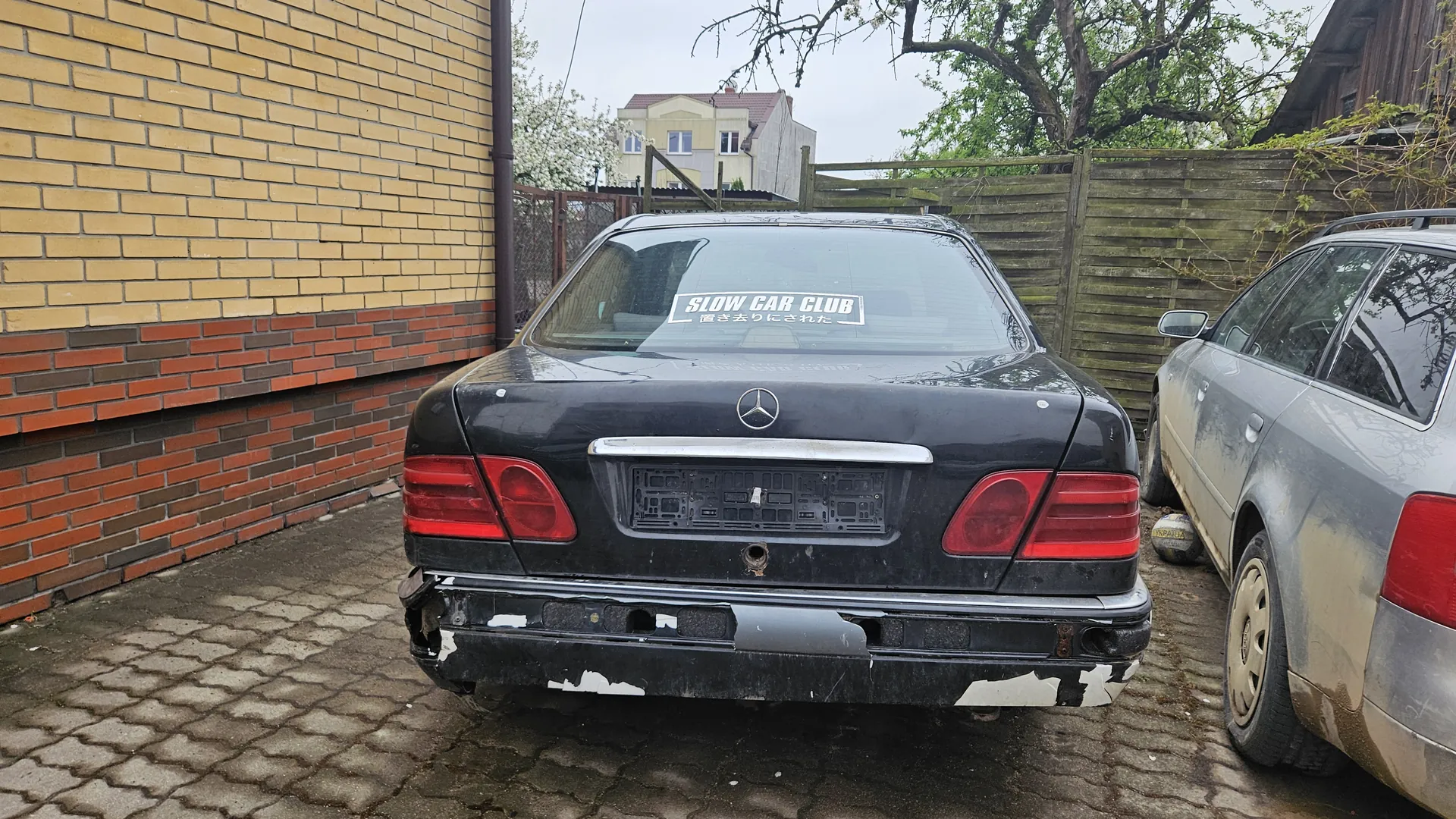 Zdjęcie przedstawia czarny samochód marki Mercedes-Benz z tyłu, zaparkowany na brukowanej powierzchni. Samochód ma uszkodzony tylny zderzak z widocznymi białymi plamami i brakującymi elementami. Na szybie tylnej naklejka z napisem "SLOW CAR CLUB" oraz japońskimi znakami. Obok, po prawej stronie, częściowo widoczny srebrny samochód. W tle żółto-czerwona ściana z cegły, drewniane ogrodzenie, drzewa i dom z czerwonym dachem. Tablica rejestracyjna jest pusta, widoczny jedynie jej uchwyt.