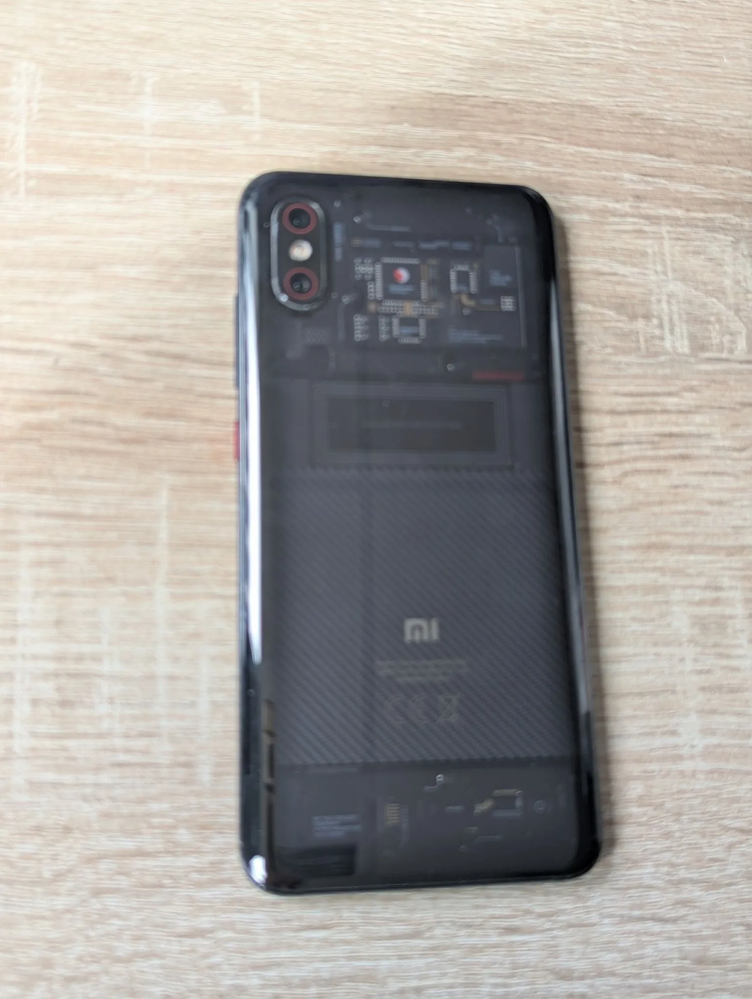 Czarny smartfon marki Xiaomi leżący na jasnym, drewnianym blacie. Telefon ma półprzezroczystą tylną obudowę, przez którą widoczne są wewnętrzne komponenty elektroniczne, w tym płyta główna i bateria. W lewym górnym rogu znajduje się moduł z dwoma aparatami fotograficznymi. Na dolnej części obudowy widoczne jest logo Mi.
