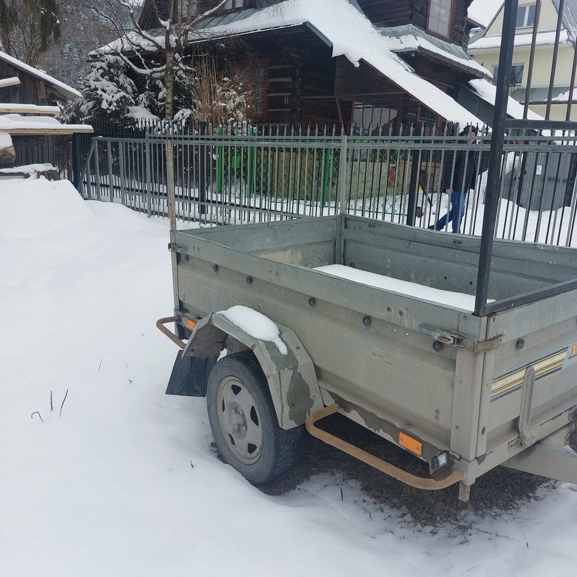 Szary przyczepa utility z śniegiem na powierzchni, zaparkowana na śnieżnej ziemi. W tle metalowe ogrodzenie z pionowymi prętami, dom z pokrytą śniegiem dachówką, drzewa z śniegiem oraz osoba w niebieskich spodniach za ogrodzeniem. Scena zimowa z obficie pokrytym śniegiem otoczeniem.