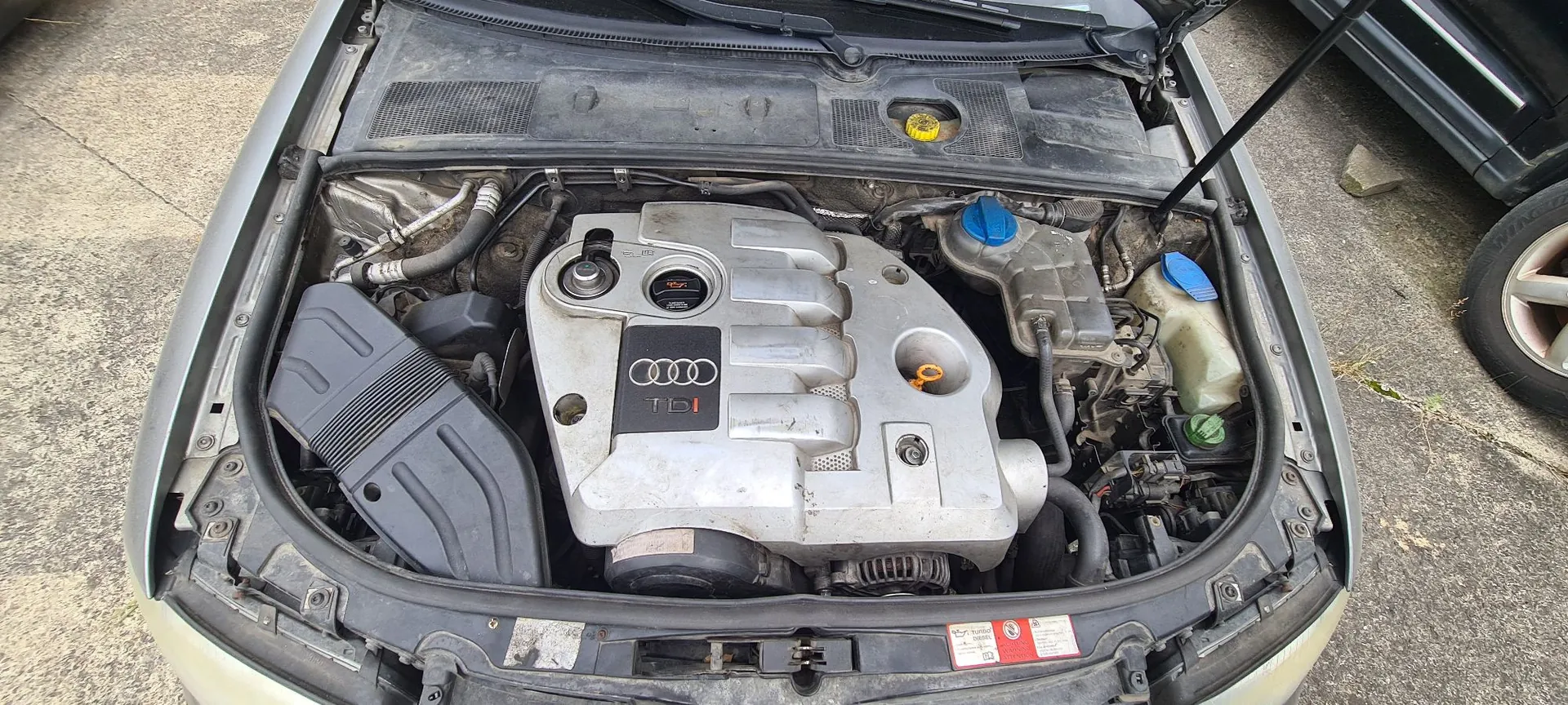 Widok z góry na otwartą maskę srebrnego samochodu, ukazujący komorę silnika. W centrum znajduje się srebrna pokrywa silnika z logo Audi i napisem TDI. Po lewej stronie widoczna jest czarna obudowa filtra powietrza, a po prawej zbiorniki z płynami eksploatacyjnymi z niebieskimi zakrętkami.