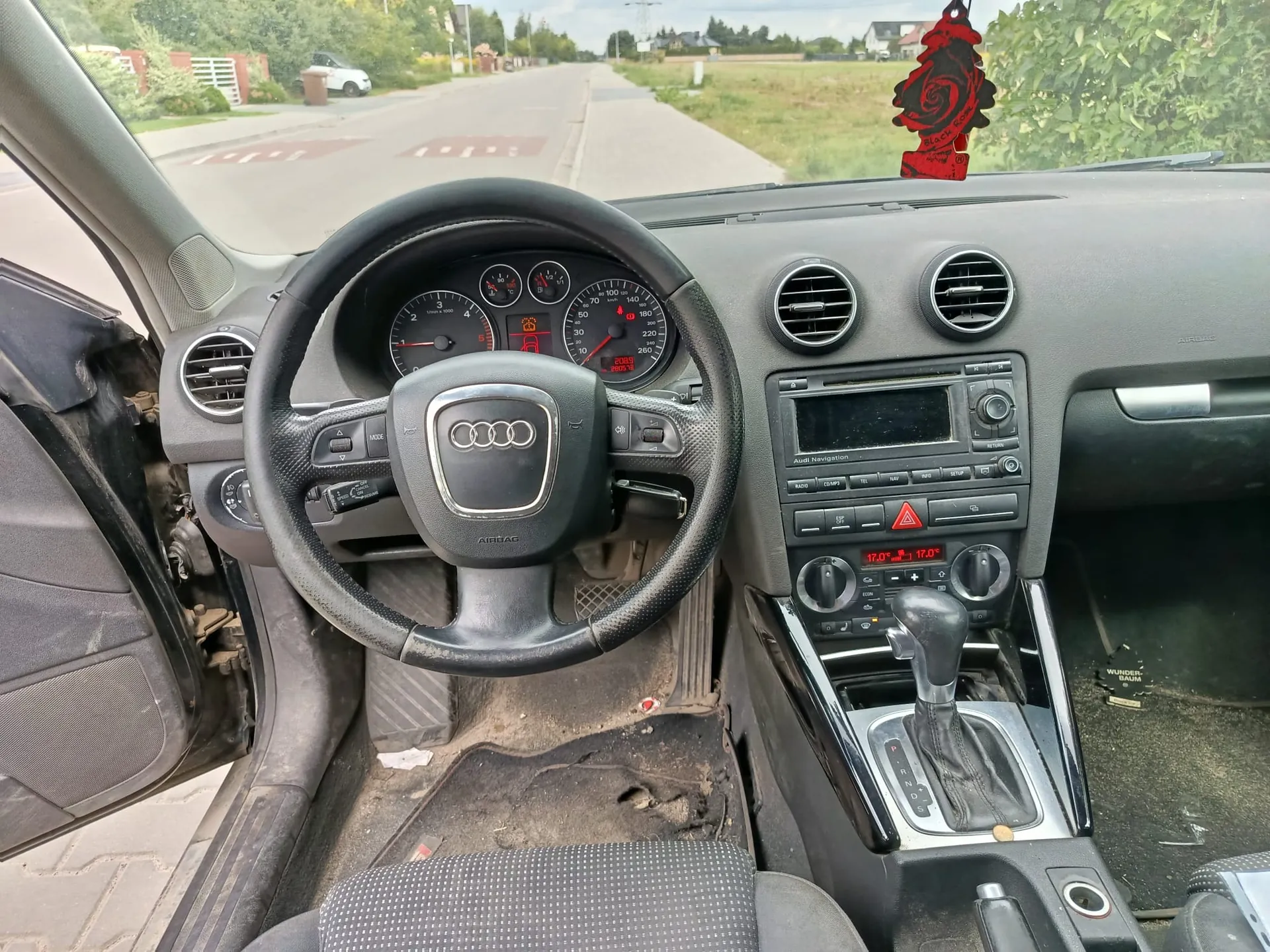 Wnętrze samochodu marki Audi widziane z perspektywy kierowcy. Widoczna jest czarna kierownica z logo Audi, deska rozdzielcza z zegarami, nawigacją oraz panelem klimatyzacji. Podłoga samochodu jest zabrudzona piaskiem i kurzem. Przez przednią szybę widać drogę, trawę i budynki w oddali. Do lusterka wstecznego przywieszona jest czerwono-czarna ozdoba.