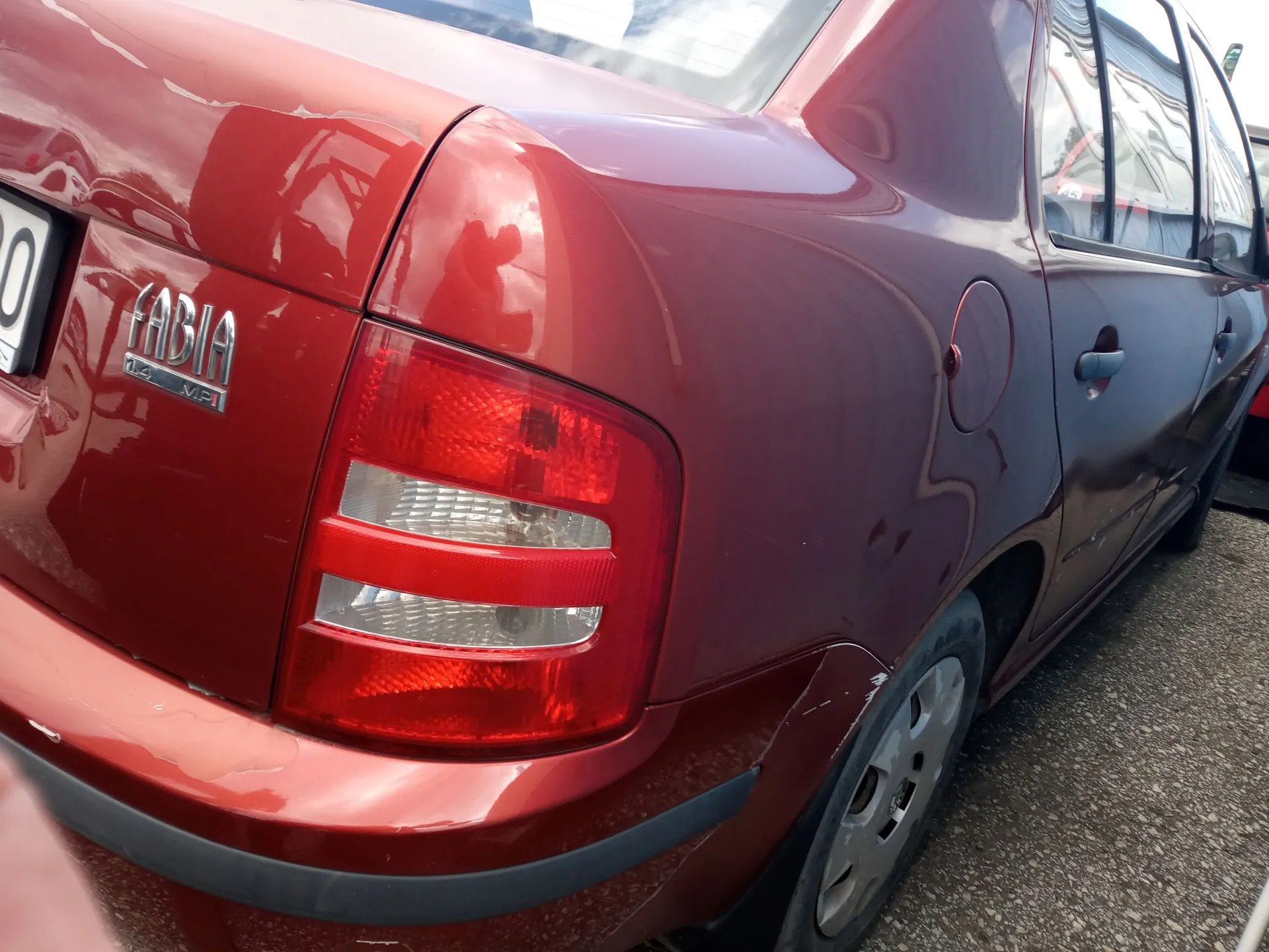 Zbliżenie na tylną część czerwonego samochodu marki Skoda. Widoczny jest napis FABIA oraz oznaczenie silnika 1.4 MPI. Na zdjęciu znajduje się tylna lampa oraz wgniecenie na tylnym błotniku nad kołem.