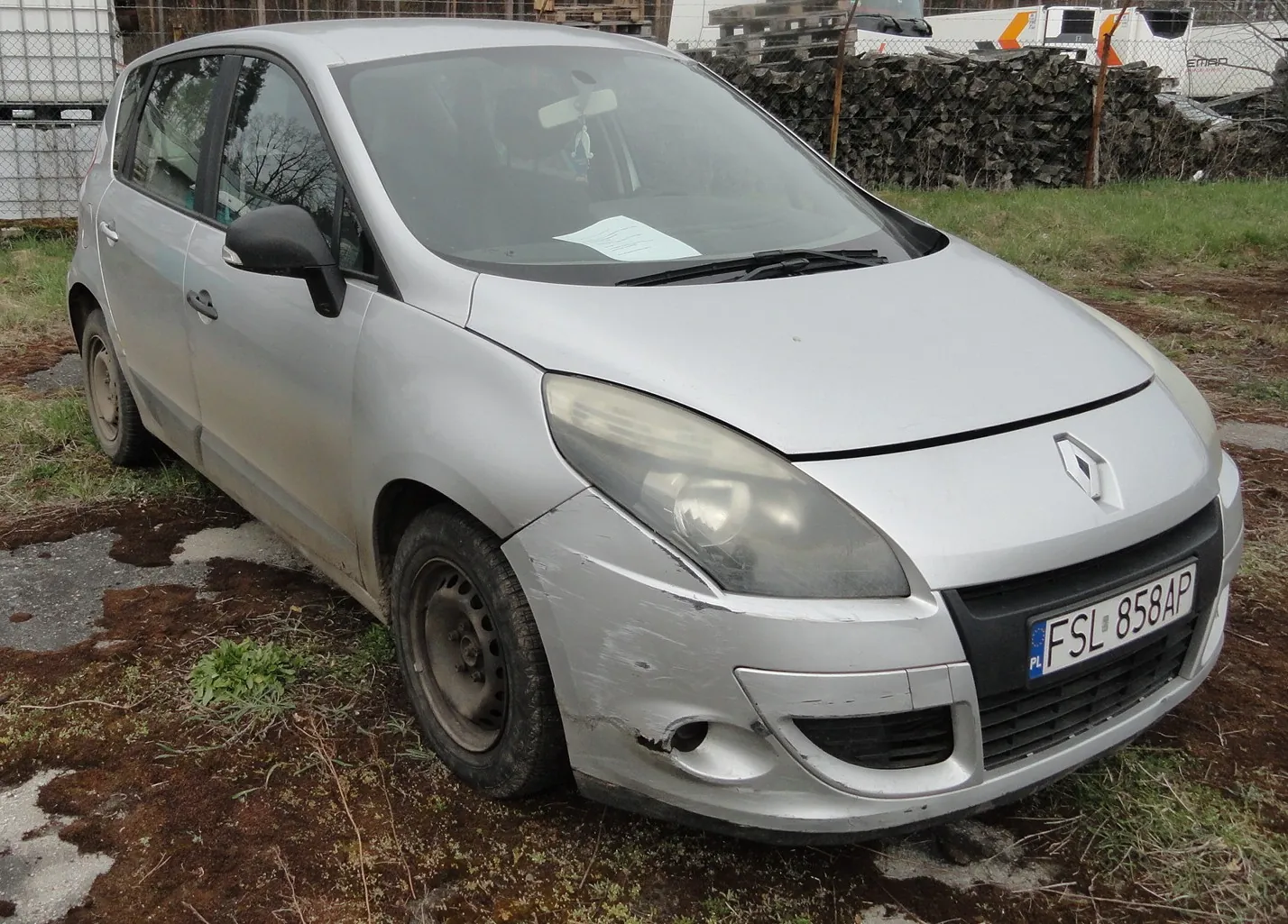 Srebrny samochód osobowy marki Renault z widocznym logo na masce, z tablicą rejestracyjną PL FSL 858AP. Pojazd ma uszkodzony przedni zderzak i brudną karoserię. Samochód stoi na nierównym terenie z trawą i odsłoniętą ziemią. W tle widoczne są stosy opon, metalowe konstrukcje oraz fragmenty innych pojazdów. Wewnątrz kabiny na desce rozdzielczej leży biały dokument.