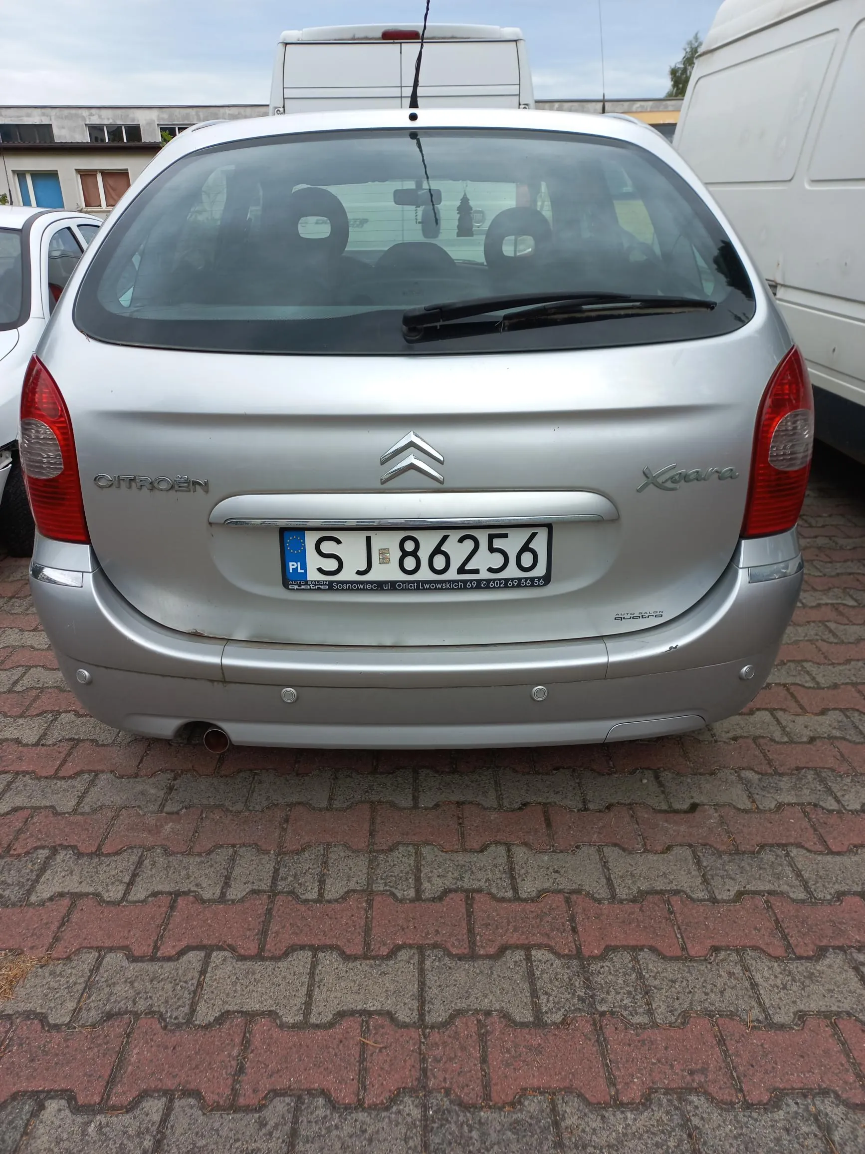 Tylny widok srebrnego samochodu marki Citroën Xsara. Na klapie bagażnika znajduje się logo marki oraz napis 'CITROËN' po lewej i 'Xsara' po prawej stronie. Samochód posiada polską tablicę rejestracyjną o numerze SJ 86256. Pojazd stoi na kostce brukowej w kolorach szarym i czerwonym.