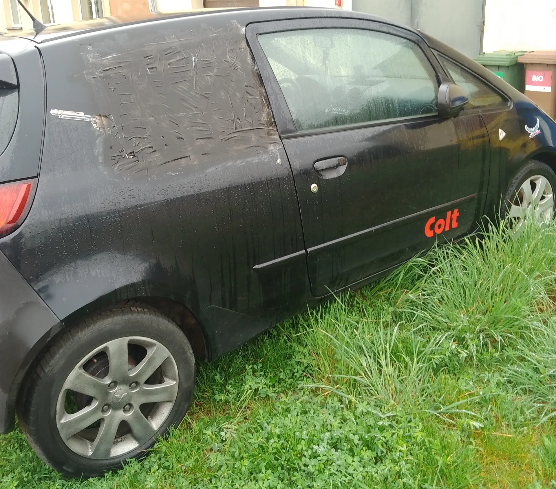 Czarny samochód osobowy typu hatchback zaparkowany na trawie. Widoczne uszkodzenie tylnego panelu bocznego z naklejonym materiałem. Na boku pojazdu czerwony napis 'Colt'. W tle zielona trawa, budynki i pojemniki na śmieci z napisem 'BIO'. Samochód pokryty kroplami wody.