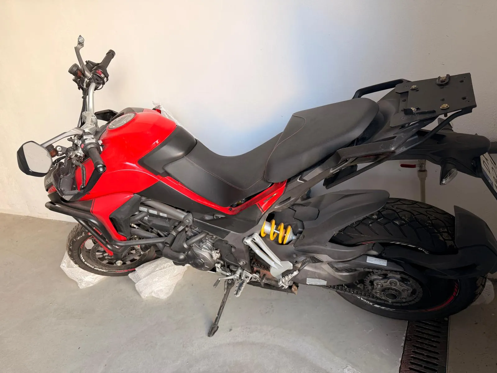 Profil motocykla w kolorze czerwonym i czarnym, zaparkowanego w pomieszczeniu o białych ścianach i szarej podłodze. Motocykl posiada czerwony bak, czarne siedzenie, czarne ramy i koła z oponami terenowymi. Widoczny jest żółty amortyzator tylny oraz lusterka boczne.