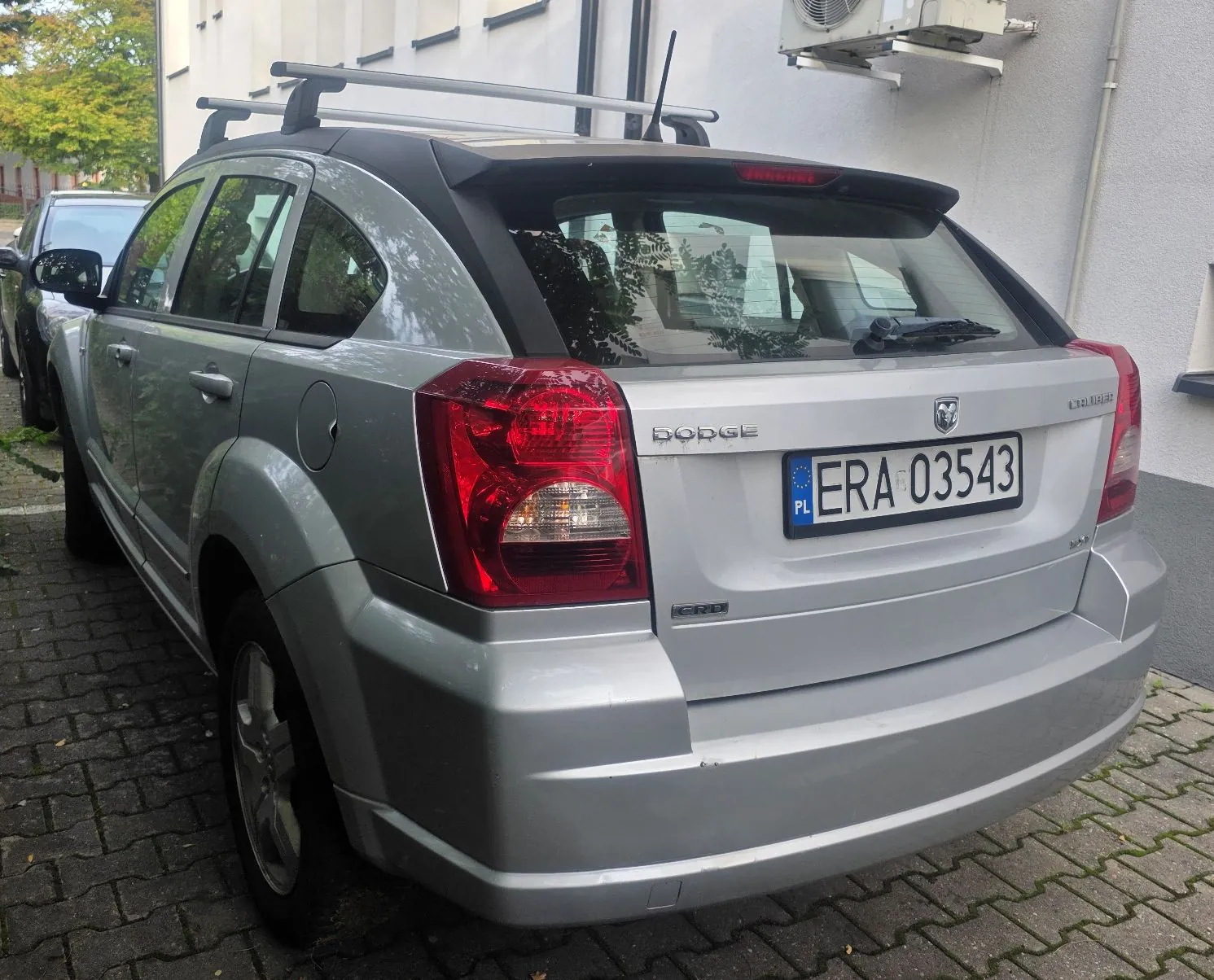 Srebrny samochód marki Dodge Caliber zaparkowany na kostce brukowej obok białego budynku. Pojazd jest widoczny od tyłu i z boku, posiada zamontowany bagażnik dachowy. Na klapie bagażnika znajduje się polska tablica rejestracyjna o numerze ERA 03543 oraz logo marki Dodge.