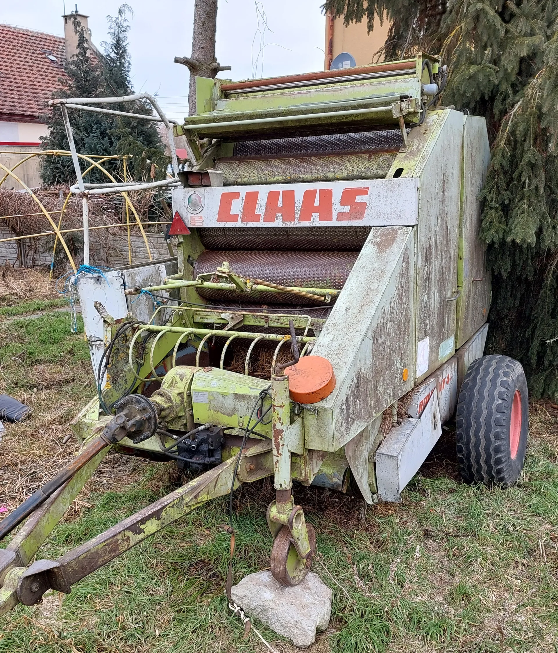 Stara, zardzewiała maszyna rolnicza marki CLAAS z zielono-białą obudową i czerwonym napisem CLAAS na przedniej części. Maszyna stoi na trawie, jedno z kół oparte jest na kamieniu. W tle widoczne są domy z czerwonym dachem, drzewa iglaste, ogrodzenie z suchymi pnączami oraz niebo. Na maszynie znajdują się metalowe elementy, przewody i pomarańczowy okrągły element.