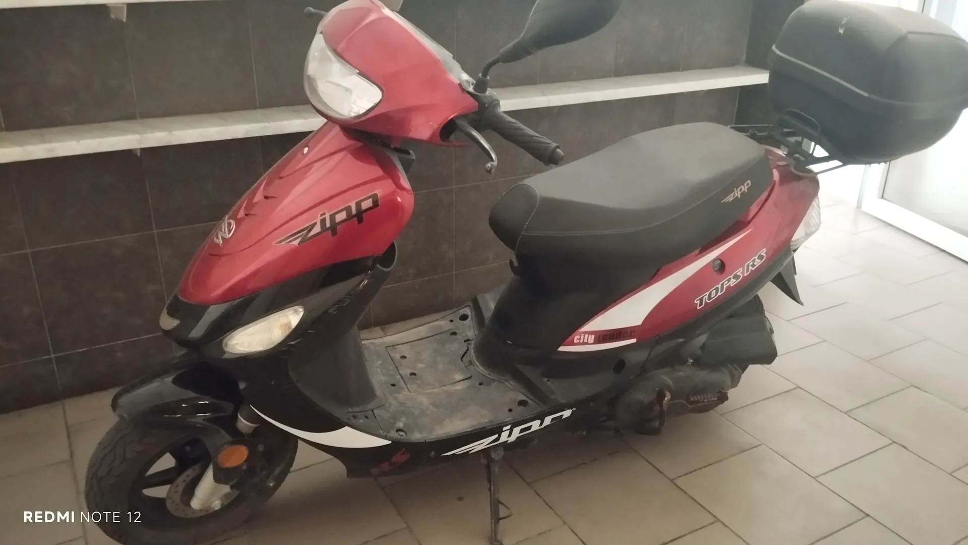 Czerwono-czarny skuter marki Zipp zaparkowany w pomieszczeniu na podłodze z jasnych płytek. Na skuterze widoczne napisy: 'Zipp' na przodzie i boku, 'TOPS RS' oraz 'city leader' na bocznej części. Skuter posiada czarne siedzenie, czarny bagażnik na tyle oraz reflektory. W tle ciemne płytki na ścianie z białymi półkami. W lewym dolnym rogu obrazu widoczny napis 'REDMI NOTE 12'.