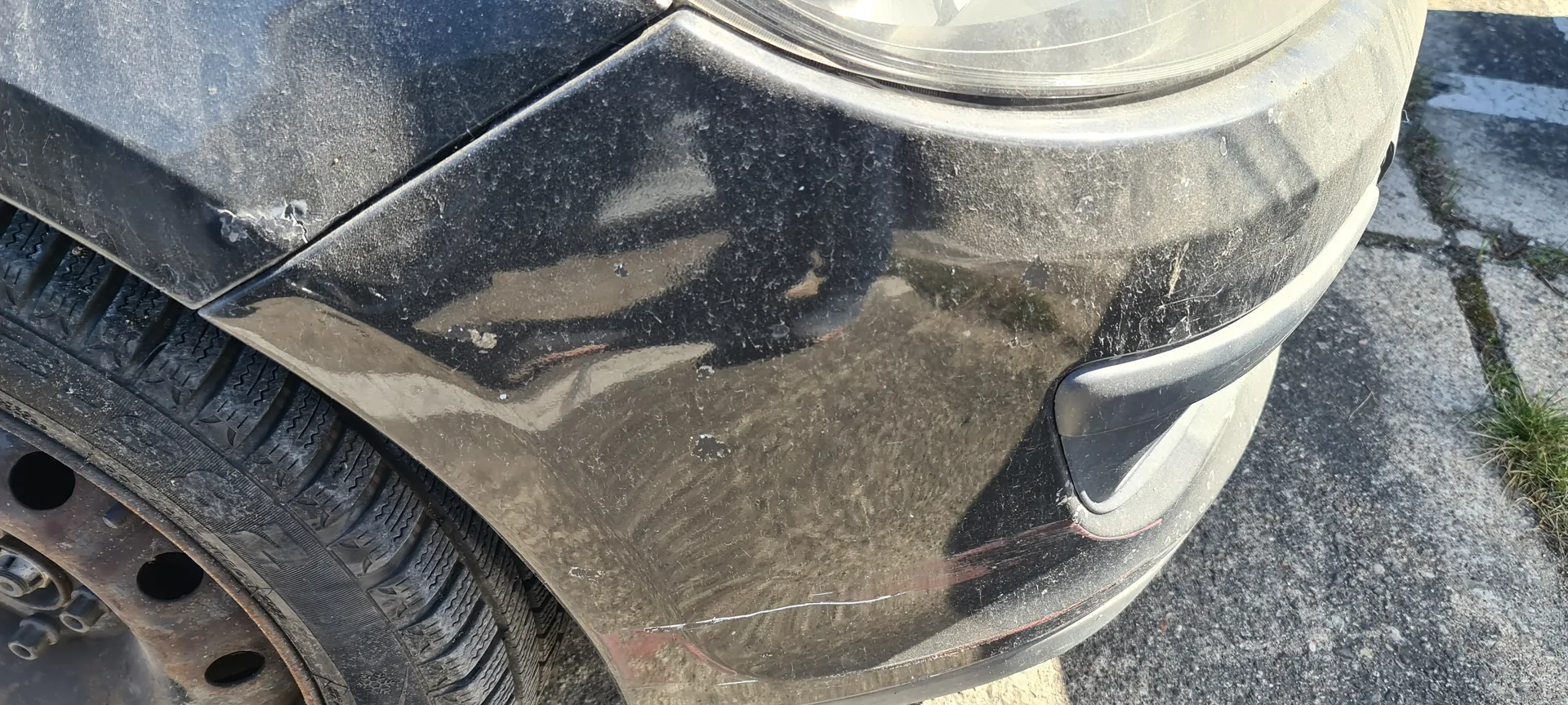 Zbliżenie na czarny samochód, widoczna przednia część z reflektorem, zarysowanym zarysem pasa przedniego i fragmentem opony. Karoseria pokryta jest kurzem i ma widoczne zadrapania. Opona z lewej strony posiada wyraźny wzór, a felga wykazuje oznaki korozji. Tło stanowi asfaltowa powierzchnia z fragmentem trawy po prawej stronie.