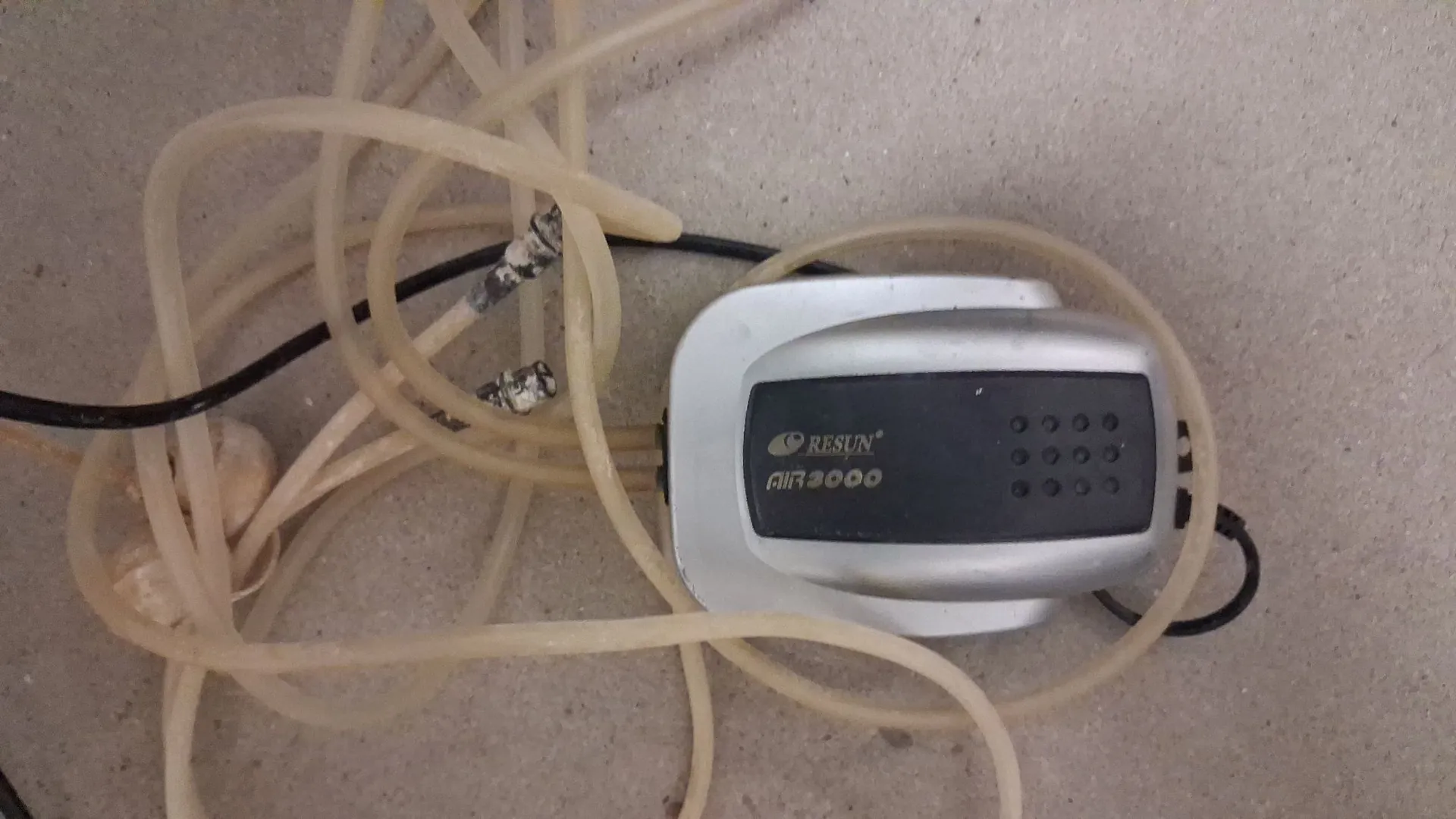 Srebrne urządzenie elektroniczne z czarnym panelem, na którym widnieje napis 'RESUN AIR 2000'. Urządzenie jest oplecione jasnymi, elastycznymi rurkami oraz czarnym kablem zasilającym.