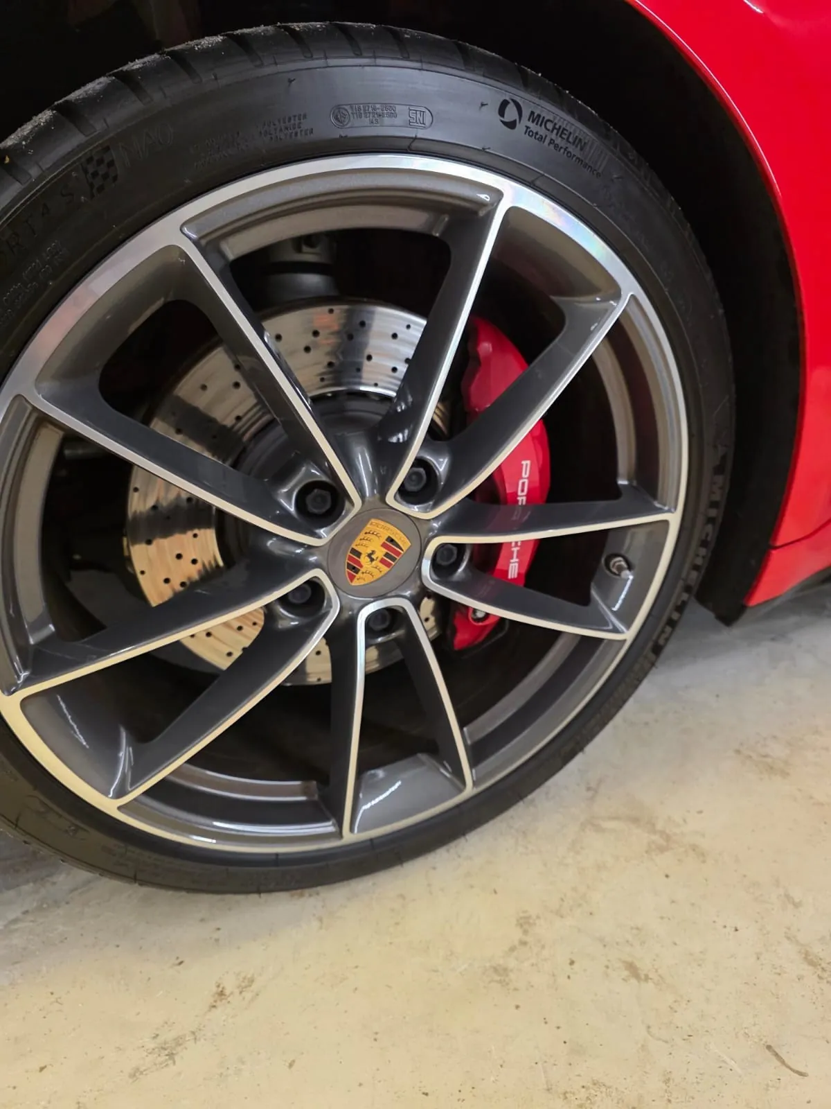Zbliżenie na koło samochodu marki Porsche. Widoczna srebrna felga z logo Porsche w środku, opona Michelin z napisem 'Total Performance' na boku, czerwony zacisk hamulcowy z napisem 'PORSCHE' oraz część czerwonego nadwozia samochodu. Tło stanowi betonowa podłoga.