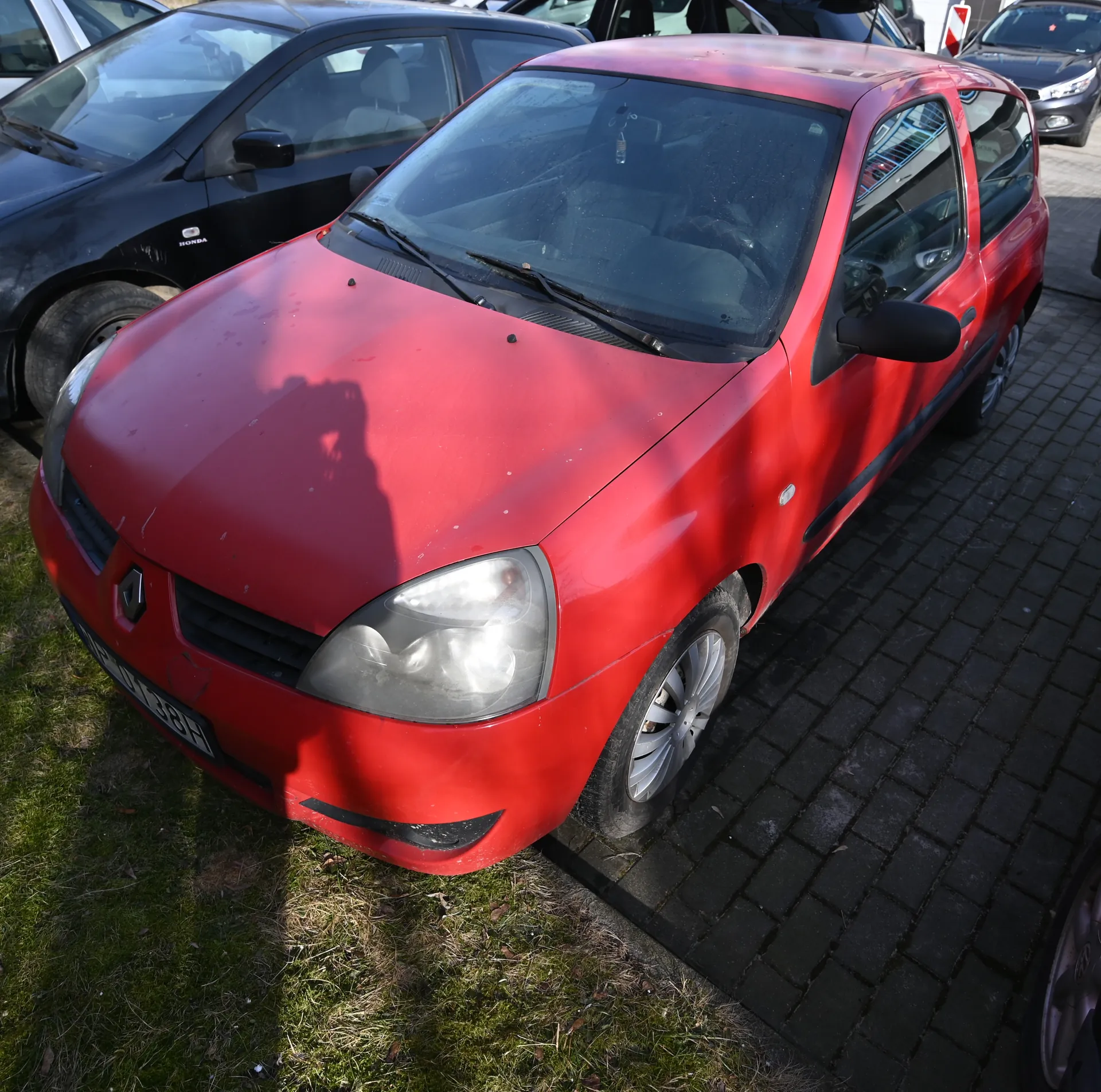 Czerwony samochód marki Renault zaparkowany na brukowanej powierzchni obok trawnika. Widoczna tablica rejestracyjna WP 0138H. W tle czarny samochód marki Honda oraz inne pojazdy. Słoneczny dzień, cień osoby fotografującej na masce czerwonego samochodu.