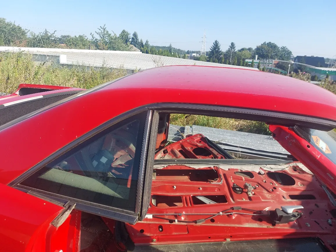 Zbliżenie na czerwony samochód z usuniętymi drzwiami i częścią nadwozia, odsłaniające wewnętrzną strukturę ramy i podwozia w kolorze czerwonym. Widoczna jest tylna boczna szyba oraz fragment dachu. W tle znajduje się trawa, biały budynek oraz drzewa pod jasnym, niebieskim niebem.