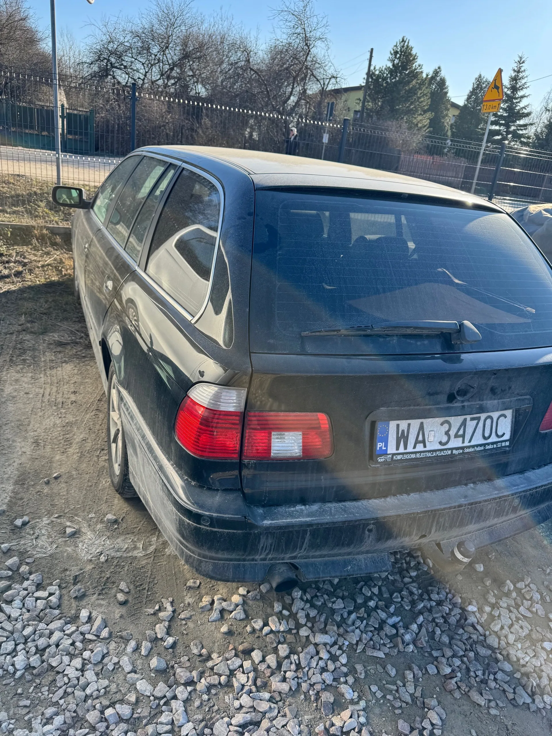 Czarny samochód typu kombi widziany od tyłu, zaparkowany na podłożu z drobnych kamieni i ziemi. Widoczna tablica rejestracyjna z napisem WA 3470C oraz oznaczeniem PL. W tle metalowe ogrodzenie, suche drzewa, drogowskaz z żółtym trójkątem i sylwetką jelenia oraz napisem 3.0 km. Jasne niebo i budynki w oddali.