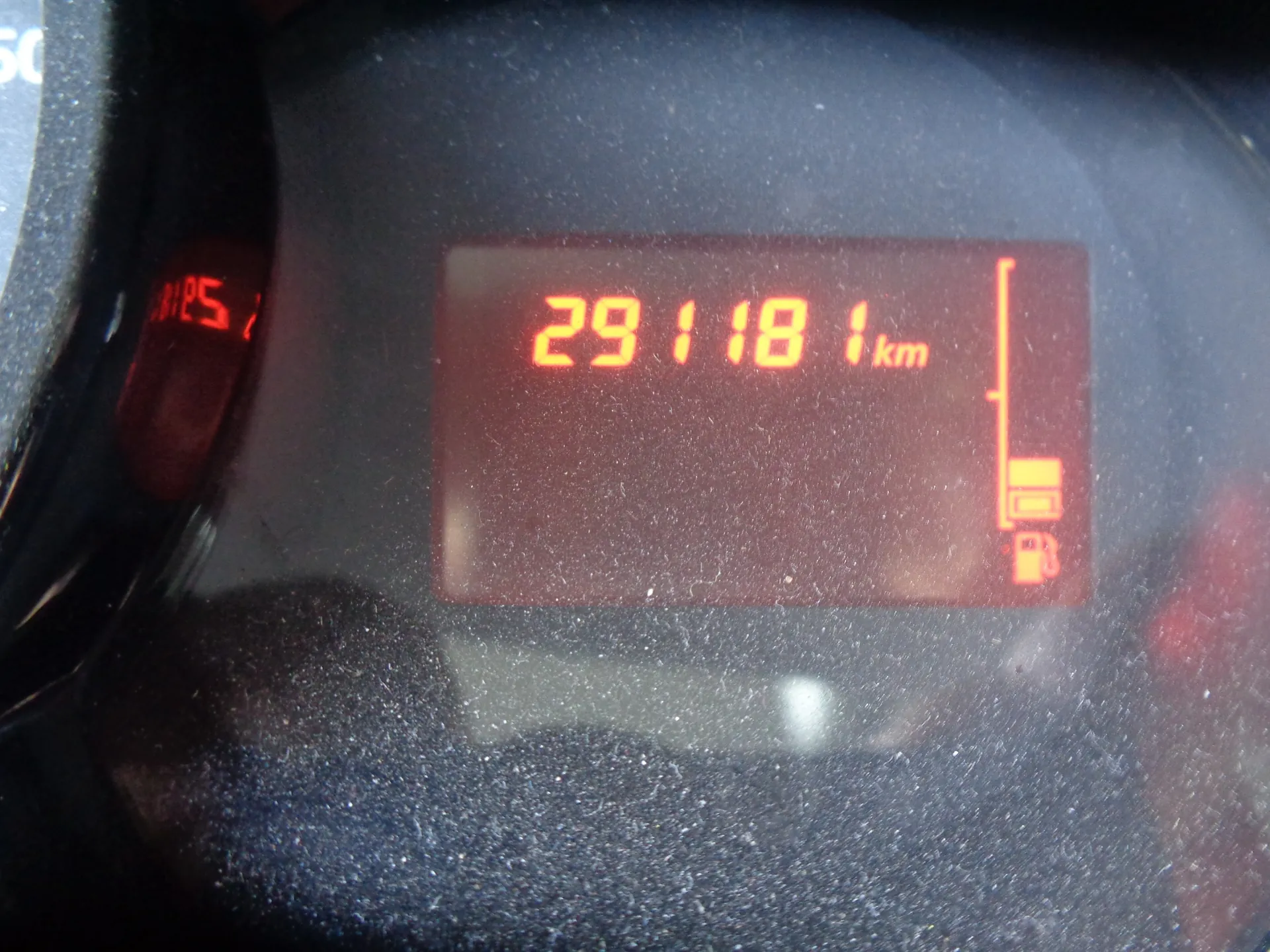 Zbliżenie na cyfrowy licznik przebiegu w pojeździe. Na czerwonym wyświetlaczu widnieje liczba 29118 oraz jednostka km. Po prawej stronie znajduje się pionowa kreska z ikoną stacji paliw na dole.