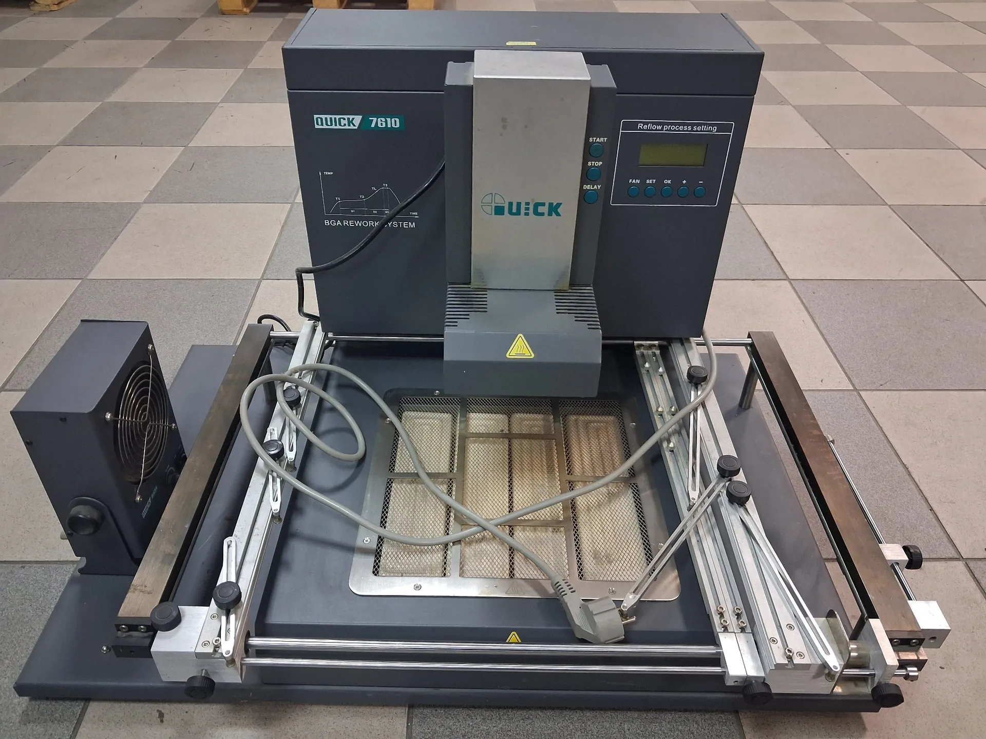 Szara maszyna do naprawy układów BGA marki QUICK, model 7610. Urządzenie posiada panel sterowania z wyświetlaczem i przyciskami, napis 'QUICK 7610' oraz 'BGA REWORK SYSTEM'. W centralnej części znajduje się ruchoma głowica grzewcza nad metalową platformą roboczą z perforowaną powierzchnią. Maszyna jest ustawiona na szarych płytkach podłogowych, a po lewej stronie widoczny jest dodatkowy moduł z wentylatorem.