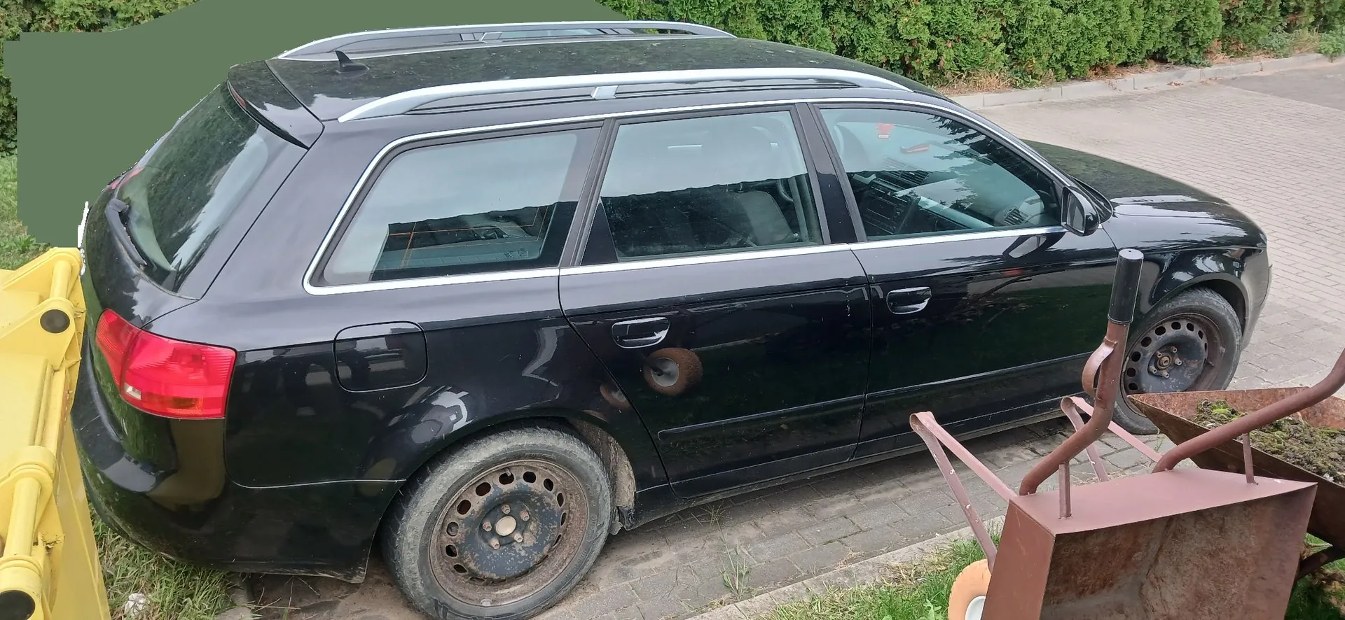 Czarny samochód typu kombi zaparkowany na kostce brukowej. Pojazd jest brudny, z widoczną korozją na nadkolu przedniego lewego koła. W prawym dolnym rogu zdjęcia widoczny jest fragment brązowej taczki, a po lewej stronie fragment żółtego urządzenia mechanicznego. W tle znajduje się zielony żywopłot.