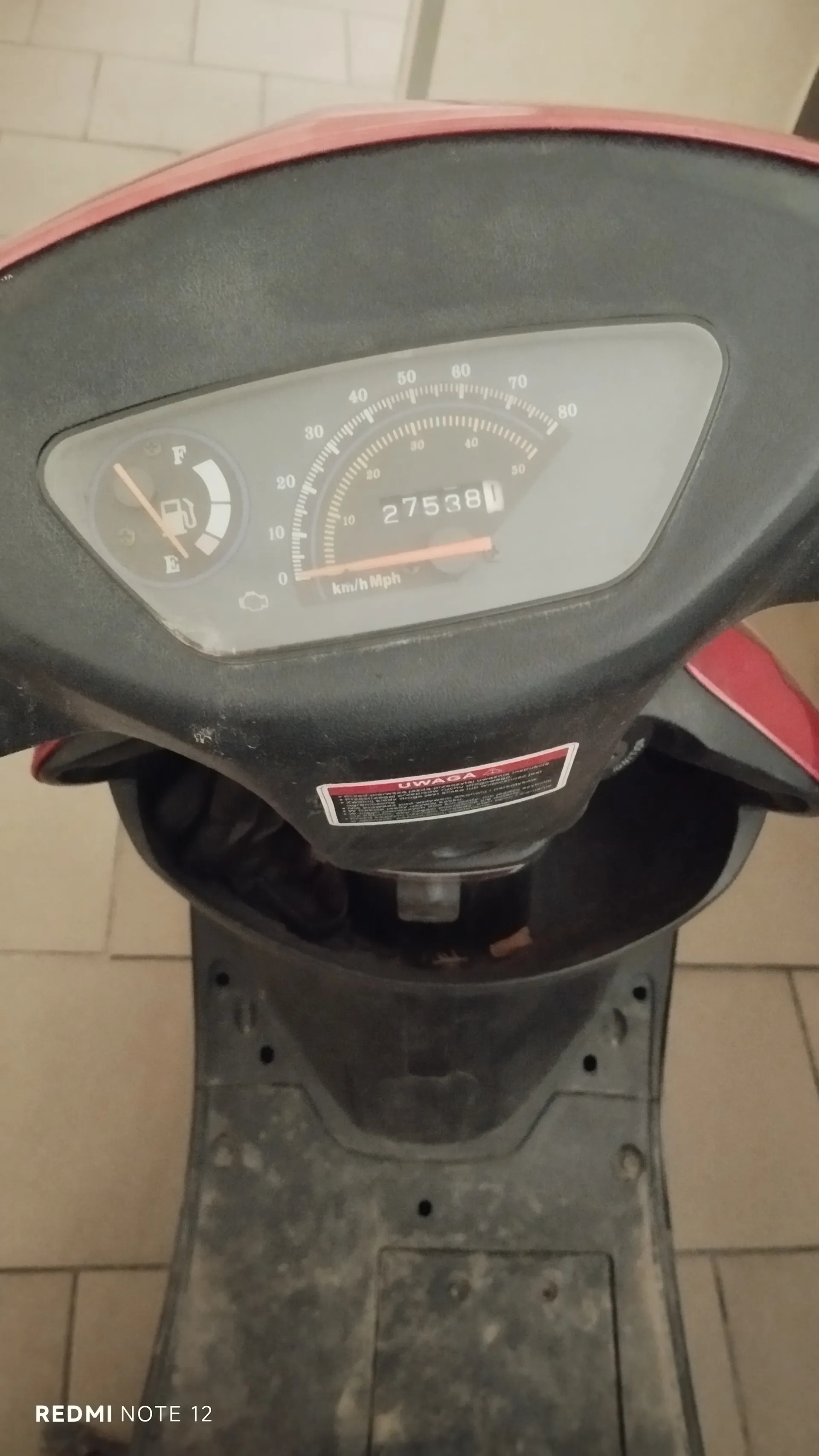 Zbliżenie na deski rozdzielcze skutera. Widoczny wskaźnik prędkości z zakresem 0-80 km/h, licznik przebiegu pokazujący 27538, wskaźnik paliwa z oznaczeniami E i F oraz ikoną zbiornika. Poniżej prędkościomierza napis 'km/h Mph'. Na desce rozdzielczej czerwono-biała naklejka z napisem 'UWAGA' oraz dodatkowym tekstem. Skuter ma czarną obudowę z czerwonymi akcentami. Tło stanowi podłoga z kafelkami. W lewym dolnym rogu widoczny napis 'REDMI NOTE 12'.