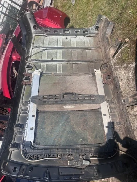 Rozłożona tylna część nadwozia samochodu z metalowym karkasem, panelami i widocznymi elementami mechanicznymi. W tle czerwony samochód, trawa i ogrodzenie. Brak czytelnego tekstu.