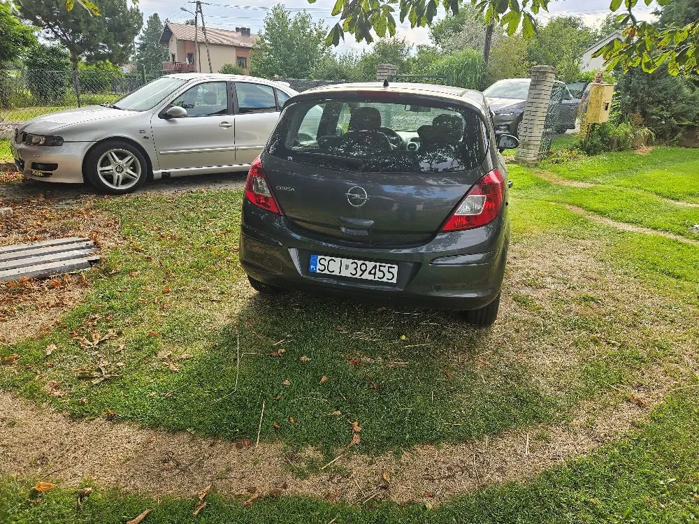 Zdjęcie przedstawiające tył ciemnoszarego samochodu marki Opel Corsa z tablicą rejestracyjną SCI 39455, zaparkowanego na trawiastym podjeździe. Obok, po lewej stronie, stoi srebrny samochód osobowy. W tle widoczne są zielone krzewy, drzewa oraz fragmenty budynków mieszkalnych.
