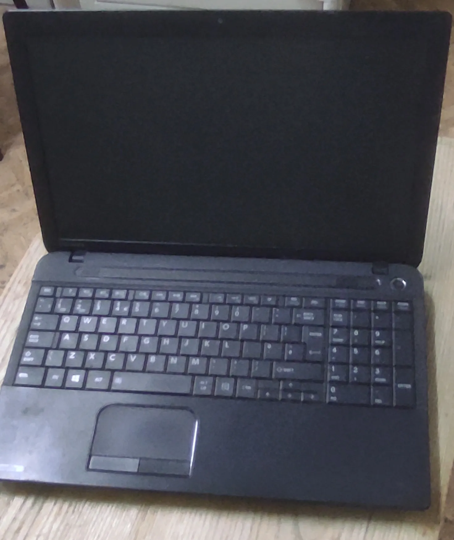 Czarny laptop z otwartym ekranem, widok z góry. Urządzenie posiada pełnowymiarową klawiaturę z blokiem numerycznym oraz touchpad na dole obudowy. Laptop spoczywa na drewnianej powierzchni.