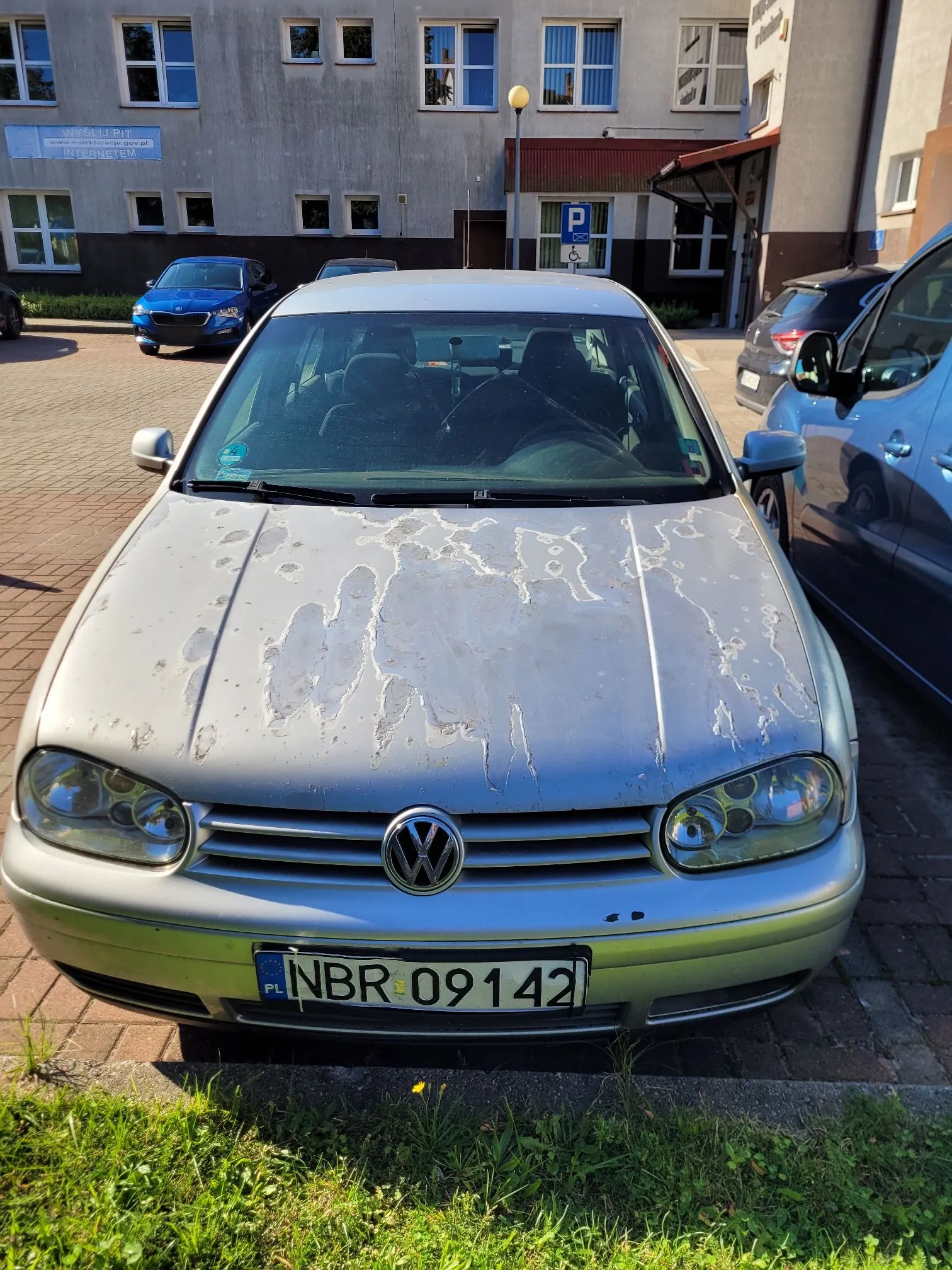 Srebrny samochód marki Volkswagen zaparkowany przodem do obiektywu. Maska samochodu jest mocno zniszczona, z widocznymi odpryskami i łuszczącym się lakierem. Pojazd posiada polską tablicę rejestracyjną o numerze NBR 09142. W tle widoczny jest szary budynek z oknami oraz inne zaparkowane samochody.