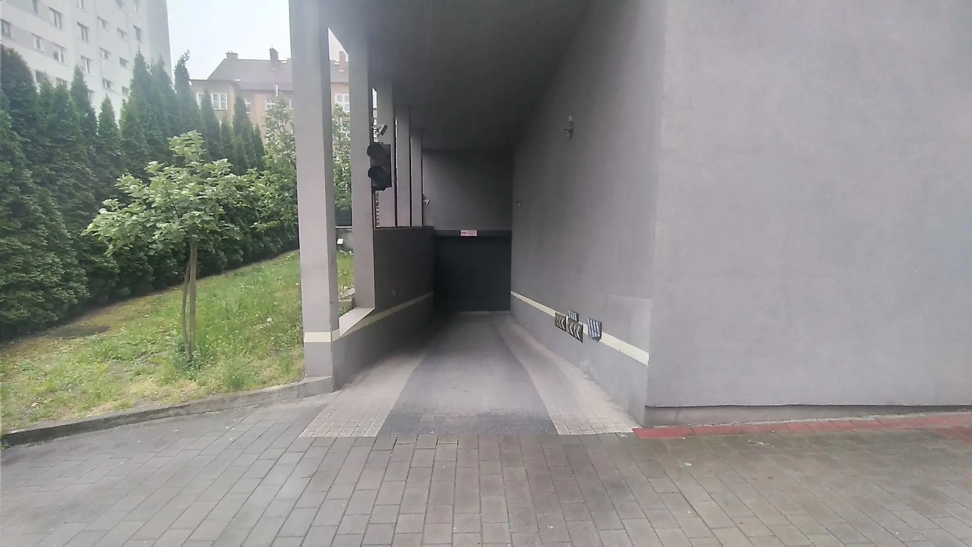 Wjazd do garażu podziemnego w nowoczesnym budynku o szarych ścianach. Podjazd prowadzi w dół w stronę ciemnego otworu wjazdu. Po lewej stronie znajduje się pas zieleni z trawnikiem i rzędem wysokich, zielonych drzew iglastych oraz jednym mniejszym drzewem liściastym. Pierwszy plan stanowi chodnik wyłożony szarą kostką brukową.