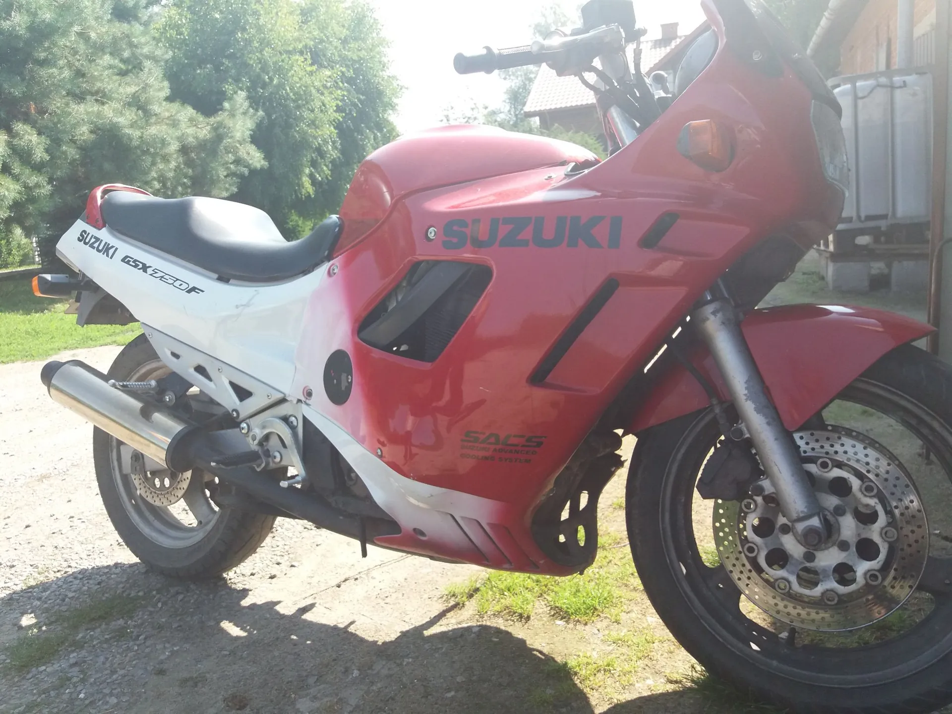 Czerwono-biały motocykl marki Suzuki, model GSX 750 F, zaparkowany na żwirowej drodze. Na czerwonej owiewce widnieje niebieski napis 'SUZUKI', a na białej części tyłu napis 'SUZUKI GSX 750 F'. Poniżej znajduje się mniejszy napis 'SACS SUZUKI ADVANCED COOLING SYSTEM'. Motocykl jest ustawiony bokiem, widoczny jest przedni widelec, koło z tarczą hamulcową oraz rura wydechowa.