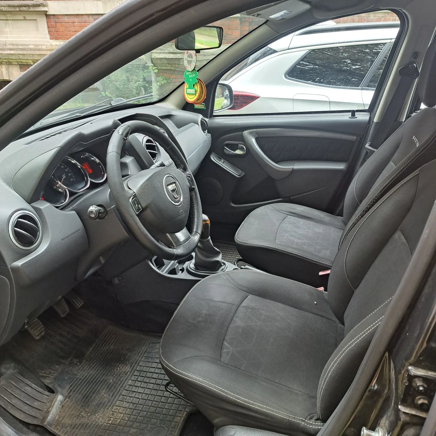 Wnętrze samochodu widziane z perspektywy otwartych drzwi pasażera. Widoczny jest czarny fotel pasażera, deska rozdzielcza, kierownica z logo Dacia, wskaźniki prędkości oraz przednia szyba, przez którą widać biały samochód zaparkowany obok.