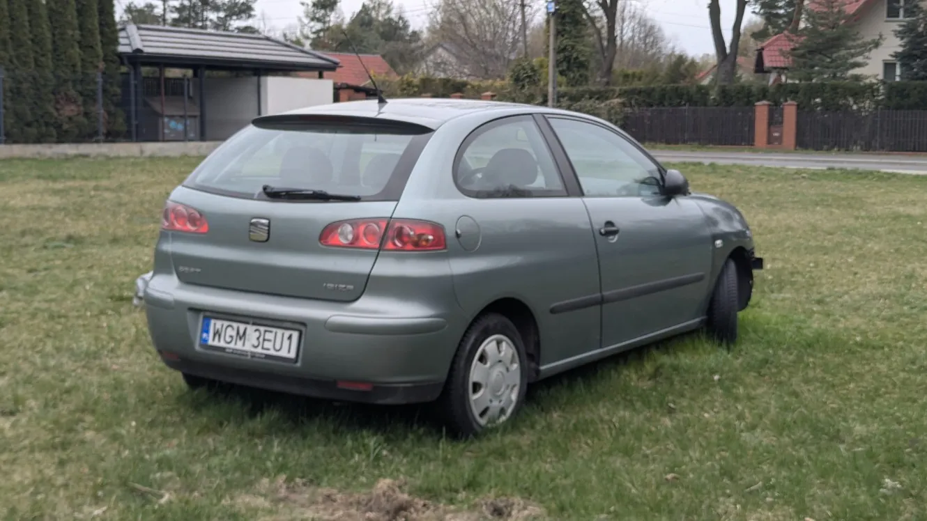 Szary samochód osobowy typu hatchback marki SEAT zaparkowany na trawie. Widoczny tył i bok pojazdu z tablicą rejestracyjną WGM 3EU1. W tle domy z czerwonymi dachami, ogrodzenie, drzewa oraz niewielka budowla z dachem. Trawa jest zielona, a na niebie widoczne są chmury.