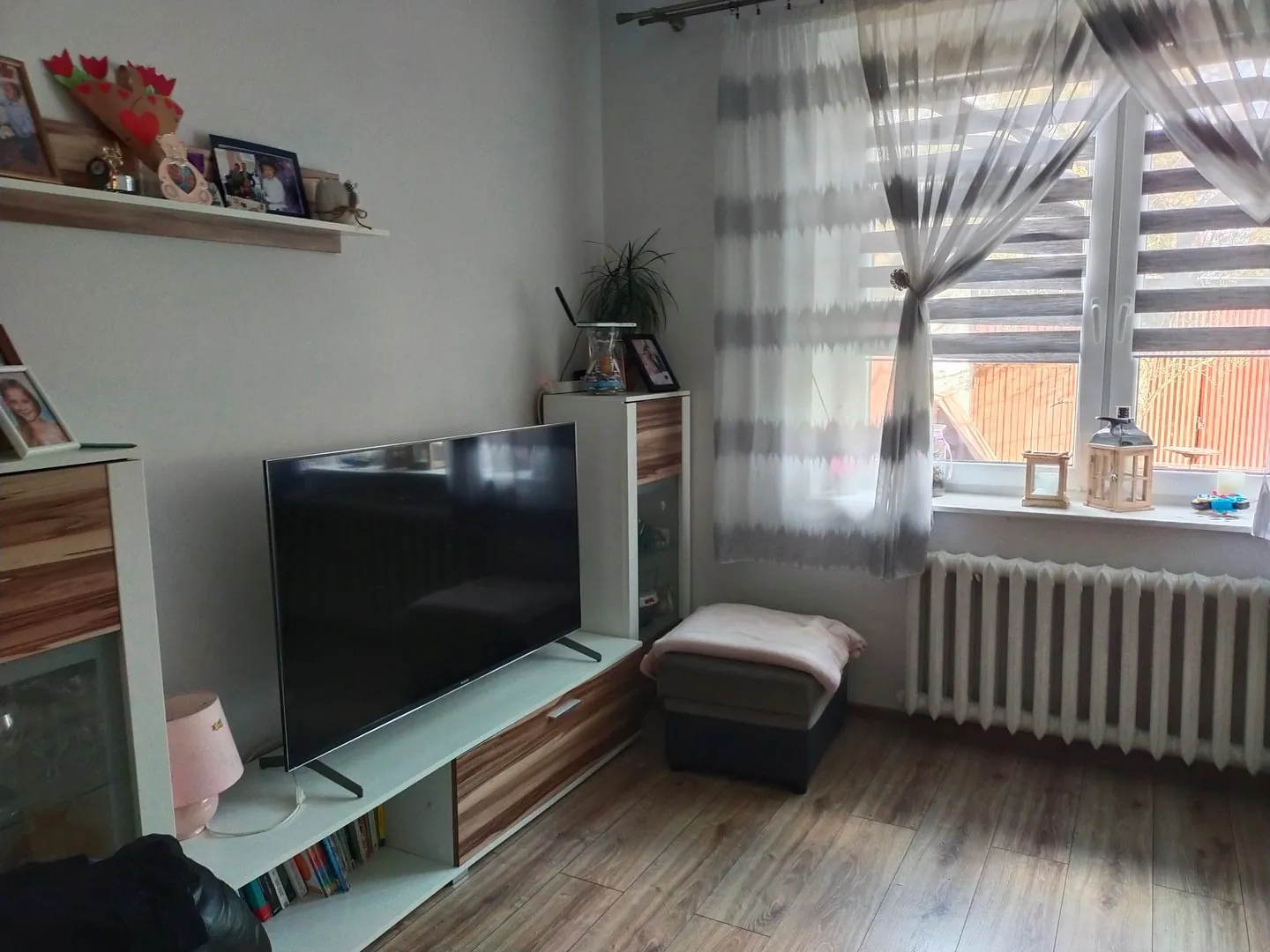 Wnętrze pokoju z telewizorem ustawionym na białej szafce RTV z drewnianymi frontami szuflad. Na szafce stoi duży, czarny telewizor z płaskim ekranem. Po lewej stronie znajduje się biała witryna, a nad telewizorem zamontowana jest biała półka z ramkami na zdjęcia i dekoracjami. Po prawej stronie stoi wysoka, wąska biała szafka. Obok niej znajduje się szary puf z jasowym kocem. W tle widoczne jest okno z białymi, półprzezroczystymi firankami w poziome pasy, pod którym zamontowany jest biały grzejnik. Podłoga wykonana jest z paneli w kolorze jasnego drewna.